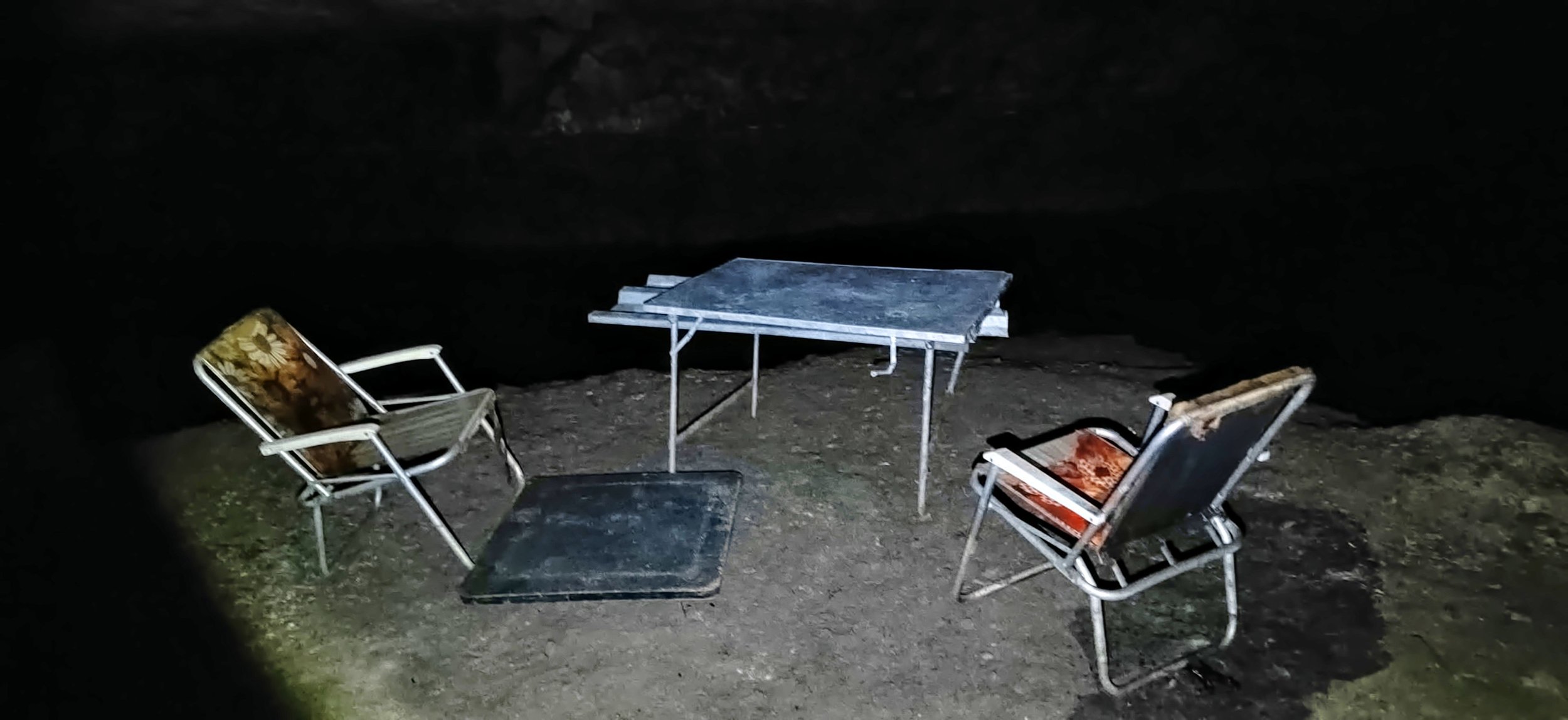 Zwei alte Gartenstühle, ein Metalltisch und ein kleiner Beistelltisch auf einem dunklen, kiesbedeckten Boden bei Nacht.