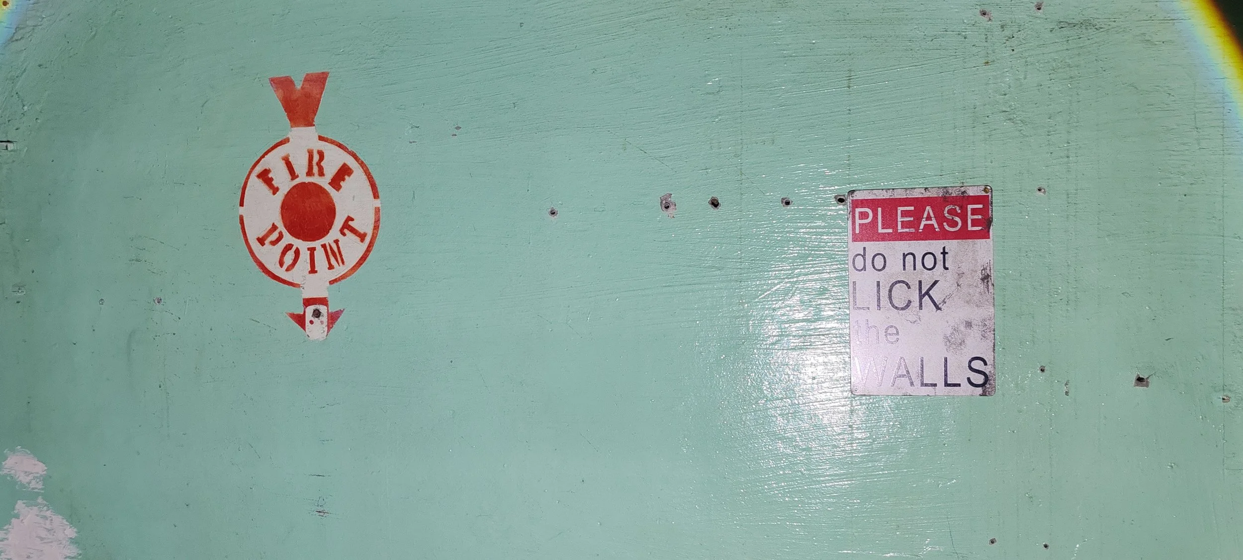 Auf einer grün gestrichenen Wand sind zwei Warnhinweise. Eine befindet sich links und zeigt ein rotes Feuer-Symbol mit der Aufschrift 'FIRE' in einem Kreis, mit Pfeilen, die nach oben und unten zeigen. Die andere befindet sich rechts und ist eine kle