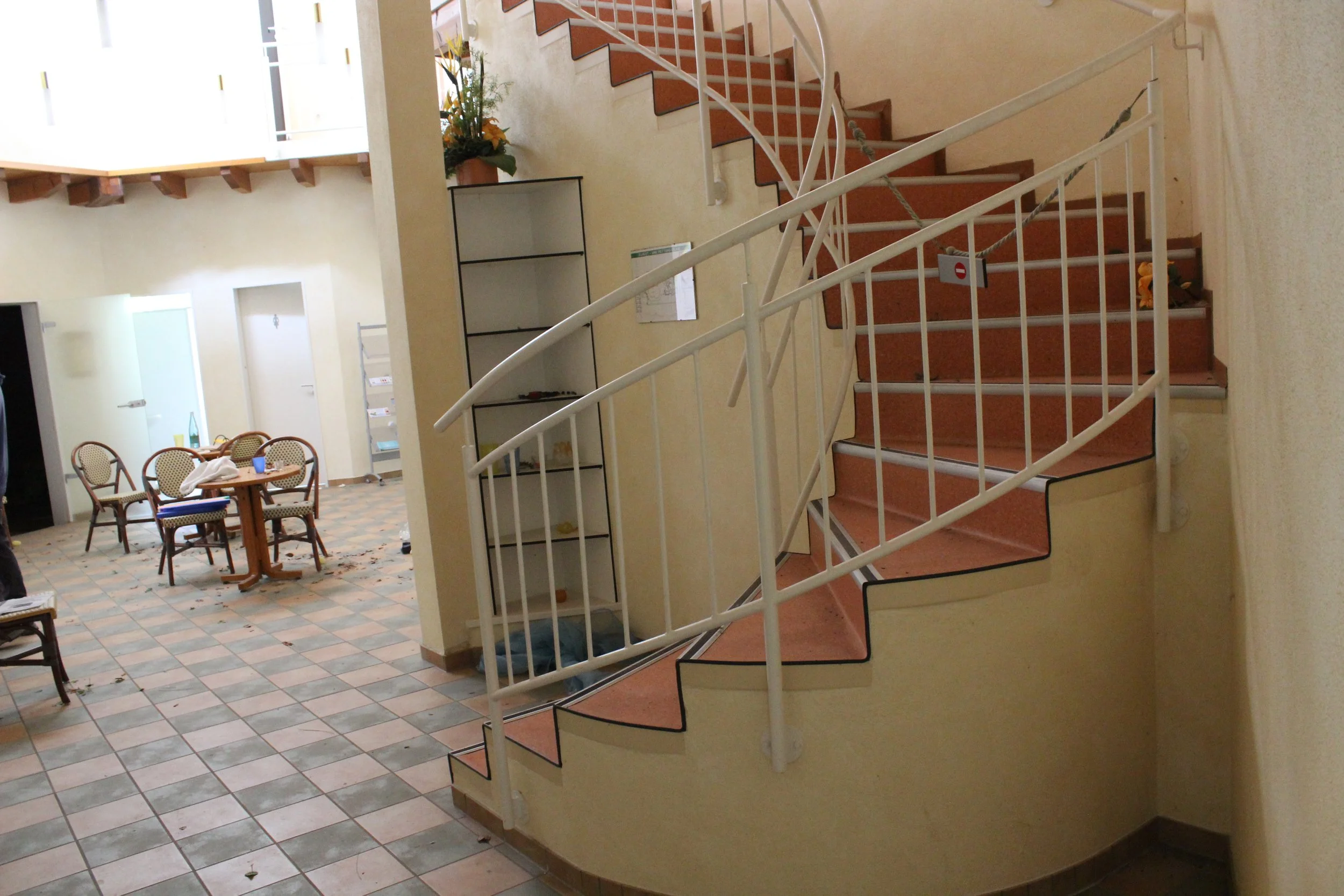 Innenraum eines Gebäudes mit Wendeltreppe, auf der einige Gegenstände liegen. Im Hintergrund stehen Tische und Stühle, die auf einem gemusterten Fliesenboden stehen.