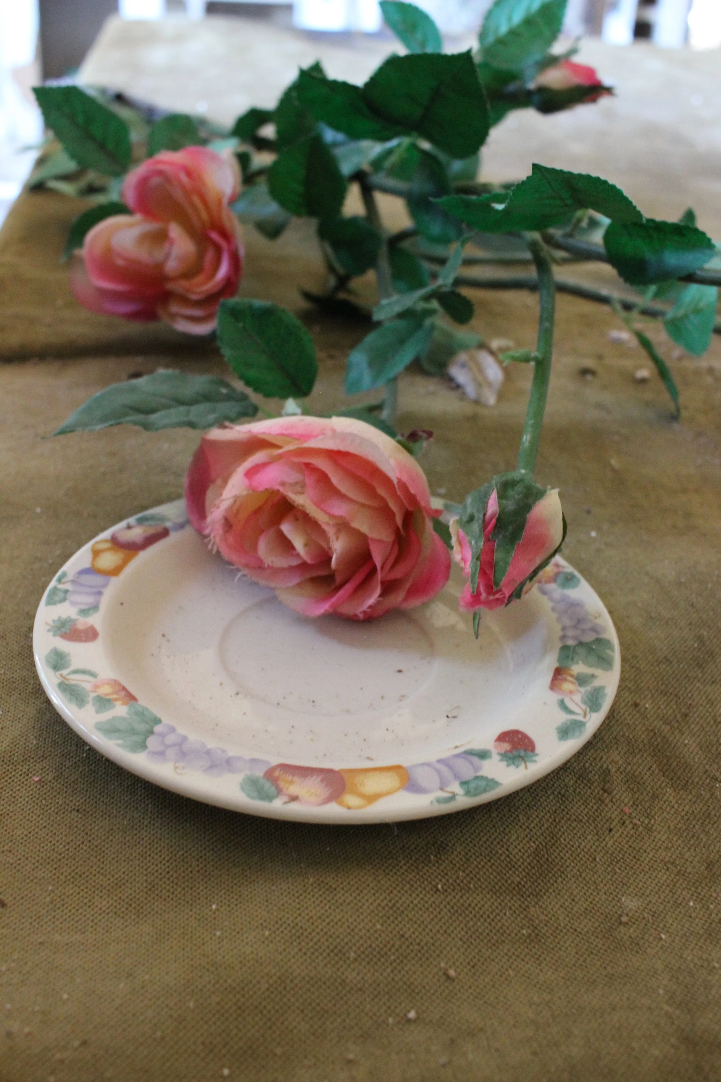 Zwei Rosen mit Blättern liegen auf einem kleinen Teller mit blumigem Muster auf einem braunen Tuch.