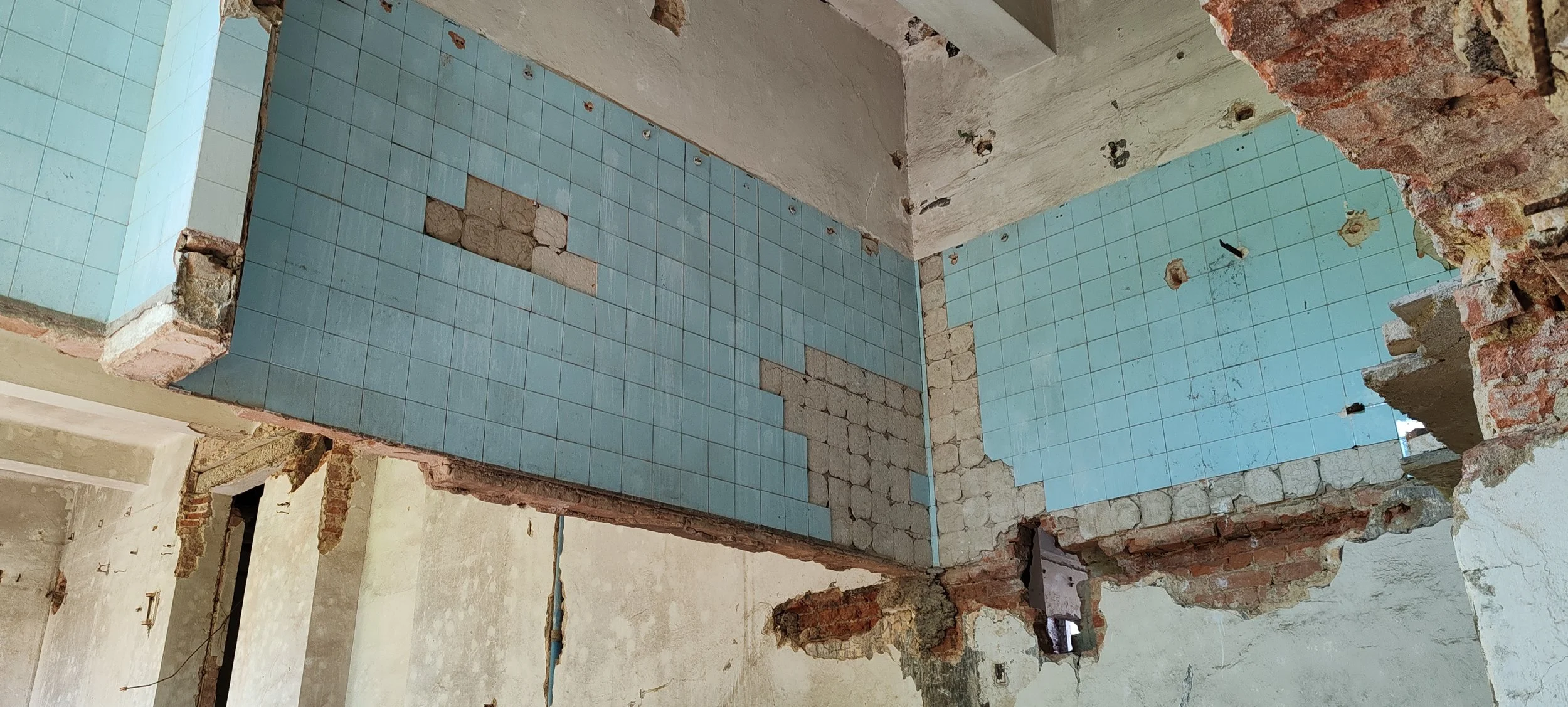 Innenansicht eines renovierungsbedürftigen Raums mit teilweise entfernter Wand, offene Ziegel und Mauerwerk sichtbar, Boden aus Fliesen und erkennbares Baumaterial an den Wänden.