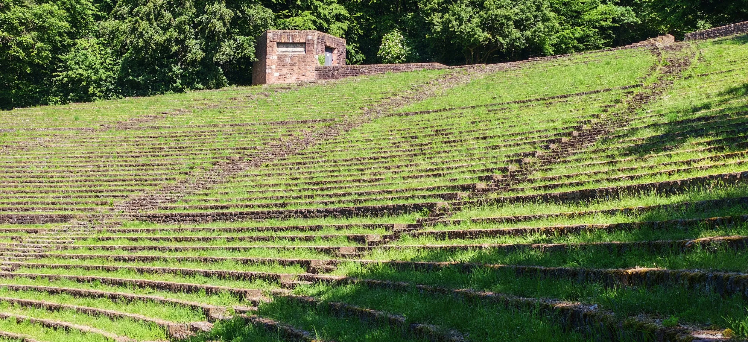 Ovale Freitreppe auf einem Hügel mit grünem Gras, im Hintergrund ein kleines Haus aus roten Steinen, umgeben von Bäumen.