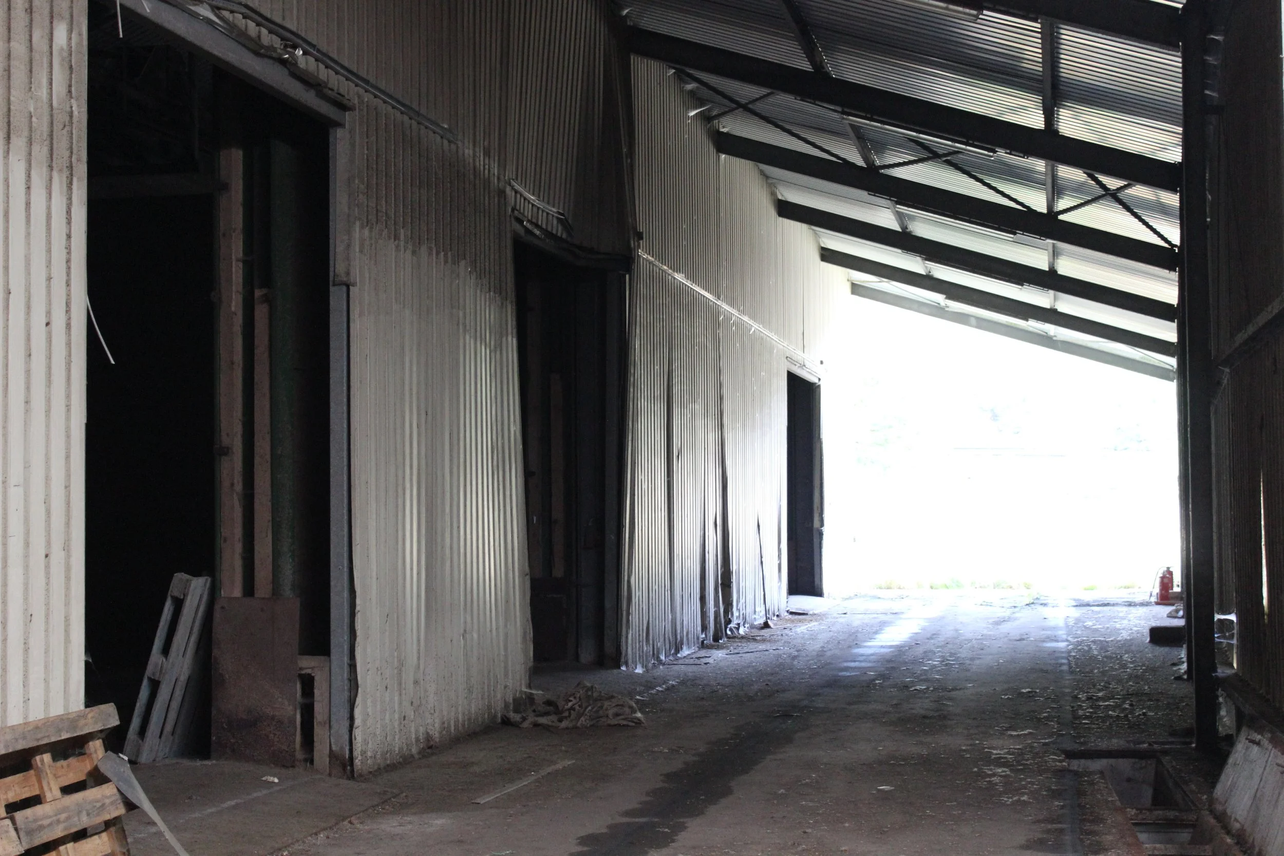 Innenansicht eines unvollständigen oder im Bau befindlichen Gebäudes mit offenem Ausgang, Wänden aus Metallverkleidung, einem Dach mit Unterkonstruktion und einer unbefestigten Bodenfläche.