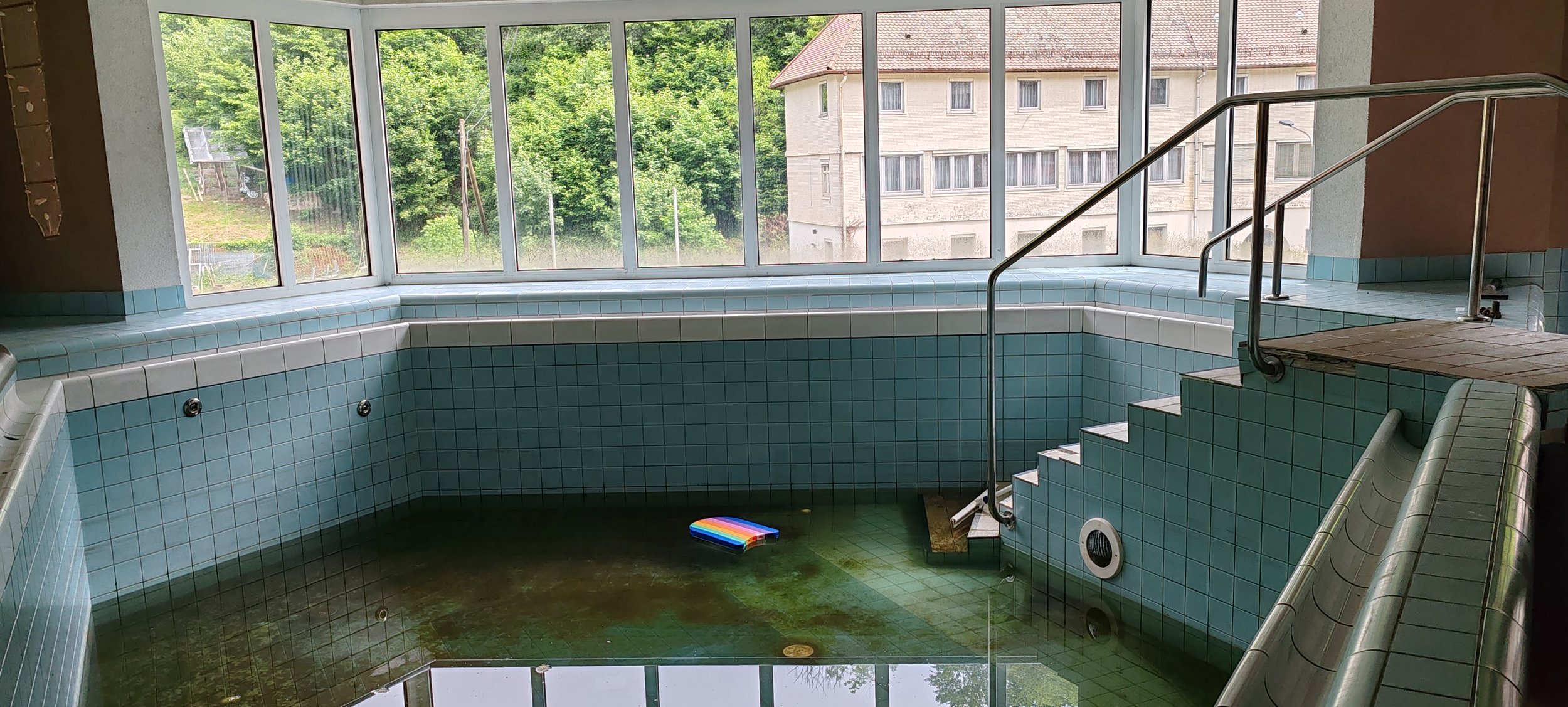 Innenraum eines alten, ungeputzten Schwimmbeckens mit grüner Bodenplatte, Wasserpfütze und einem bunten Schlauch, umgeben von großen Fenstern mit Blick auf Bäume und Gebäude.