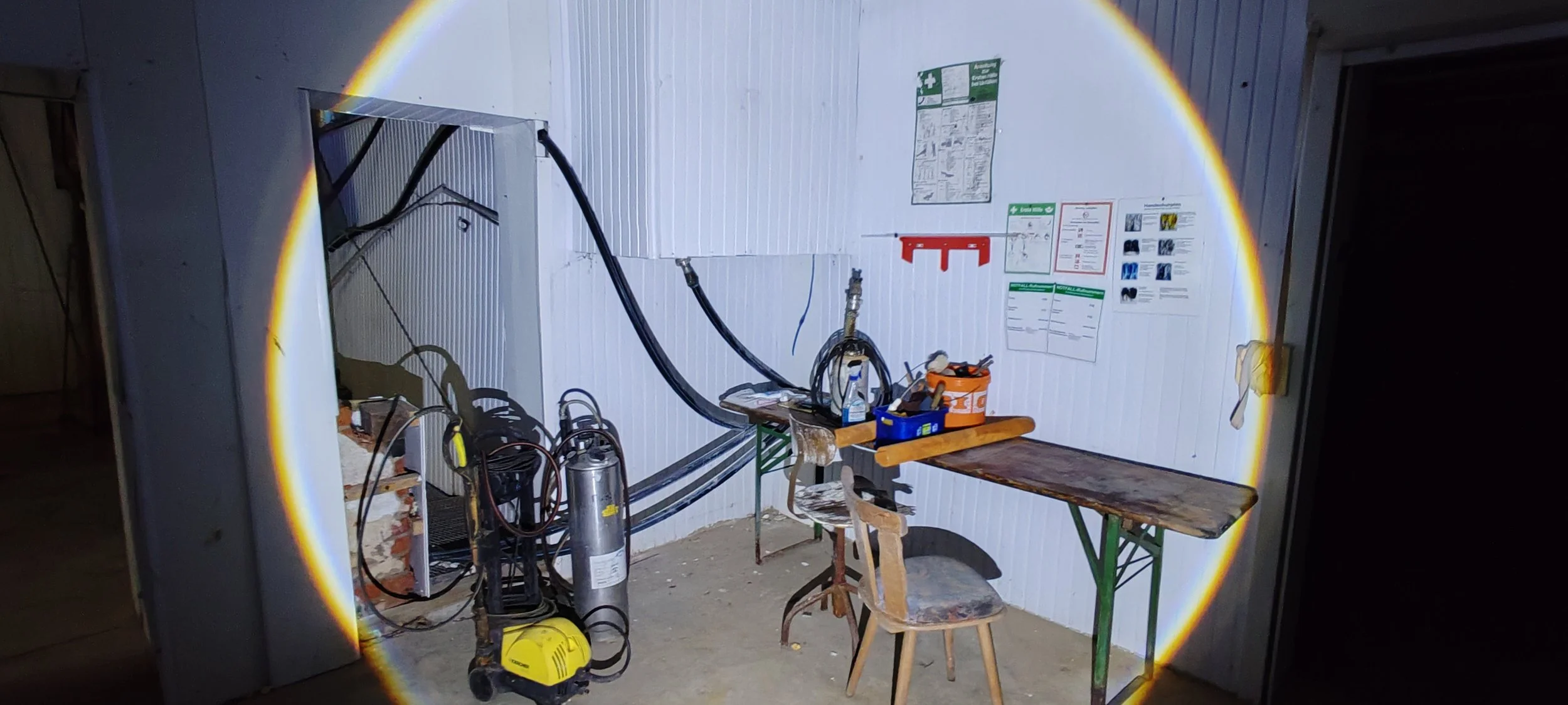 Lichtkreis beleuchtet einen kleinen Werkraum mit Werkbank, Werkzeugen, Wasserwaage, und technischen Geräten, an einer Wand mit Anleitungen und Notizen.
