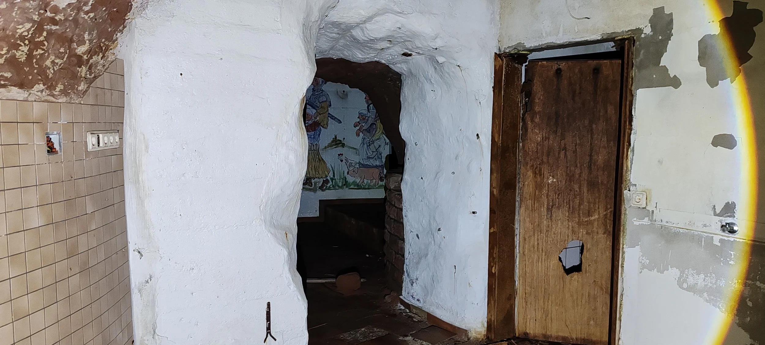 Ein alter Raum mit weißen Wänden, teilweise abgeblätterter Farbe, eine Holztür mit kleinem Loch, eine Wand mit Fliesenspiegel und eine bemalte Nische im Hintergrund.