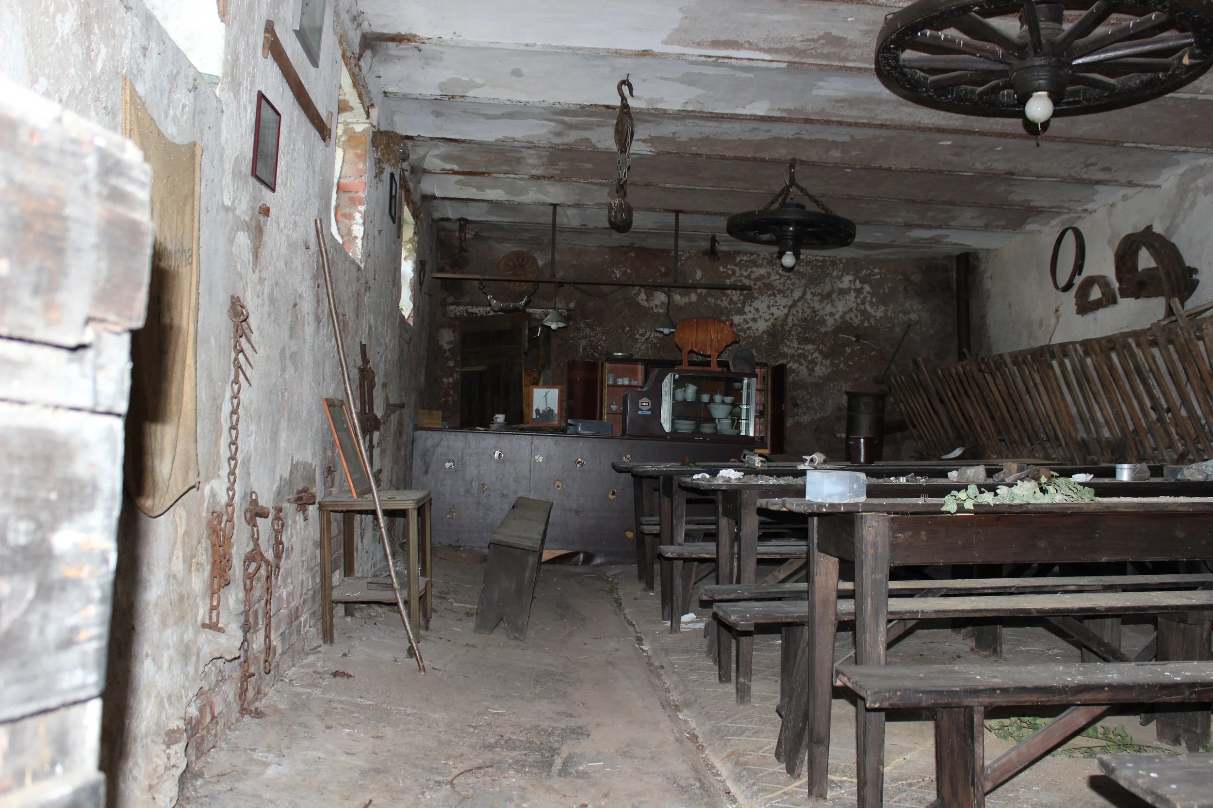 Ein verfallener, alter Raum mit alten Tischen und Stühlen aus Holz, an der Wand hängen alte landwirtschaftliche Werkzeuge, im Hintergrund ein Regal mit Geschirr, die Wände sind beschädigt und schmutzig.