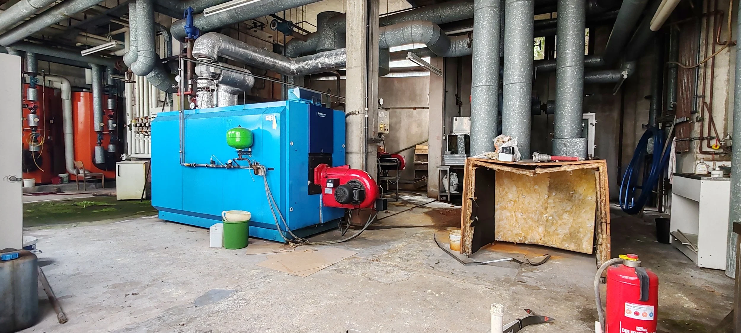 Industrielle Heizungsanlage mit blauer Maschine, rotem Ventilator, zahlreichen Rohren und Lüftungssystemen in einer Werkstatt.