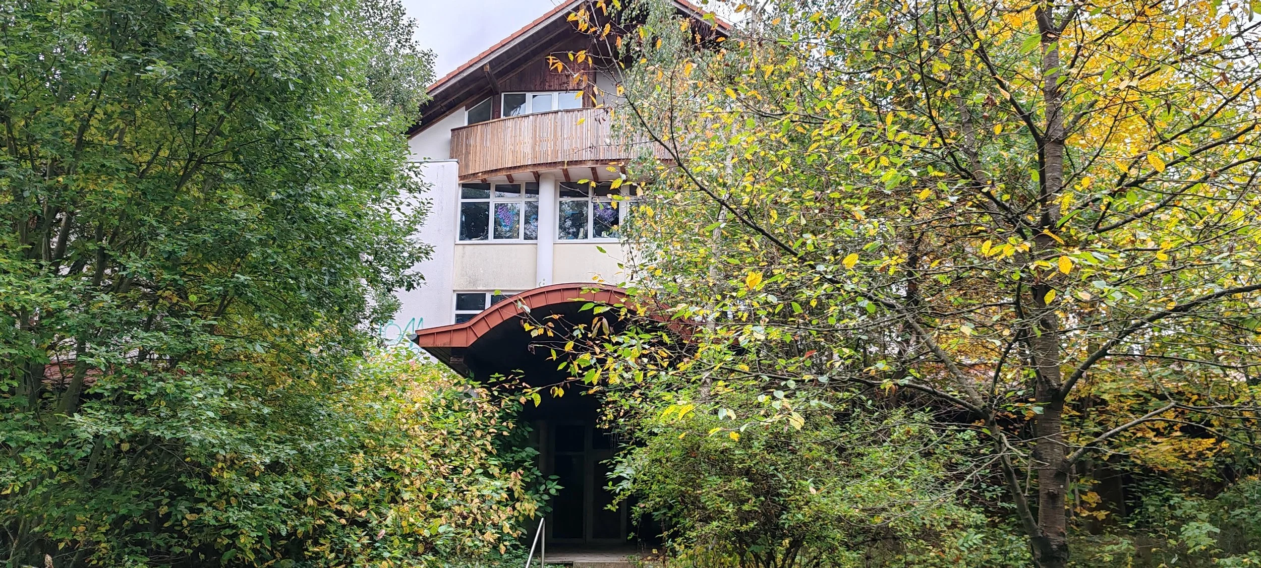 Mehrstöckiges Haus, das teilweise von Bäumen umgeben ist, mit einem roten Dach und großen Fenstern.