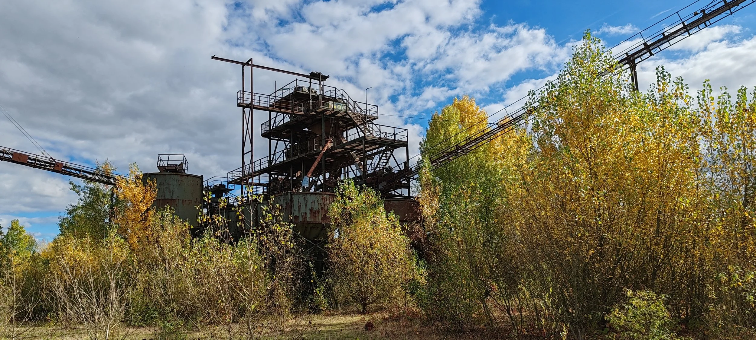Verrostete Industriemaschine im Freien, umgeben von Bäumen mit herbstlichen Blättern, unter einem bewölkten Himmel.