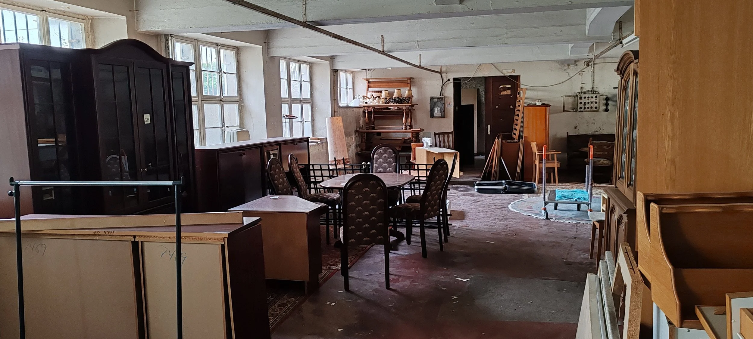 Ein Raum mit alter Möbeln, darunter Tische, Stühle, Vitrinen und Regale, teilweise auf einem neuen Teppich. Es sieht aus wie ein Lager oder ein Secondhand-Möbelgeschäft.