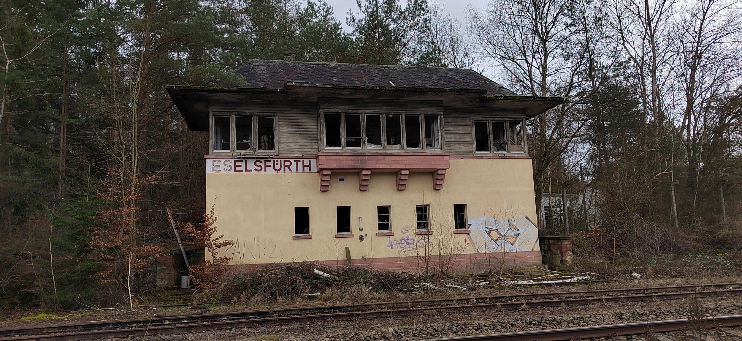 Verfallenes zweistöckiges Gebäude mit abgenutzten Fensterrahmen, Graffiti an der Wand und Bahngleise im Vordergrund, umgeben von Bäumen.