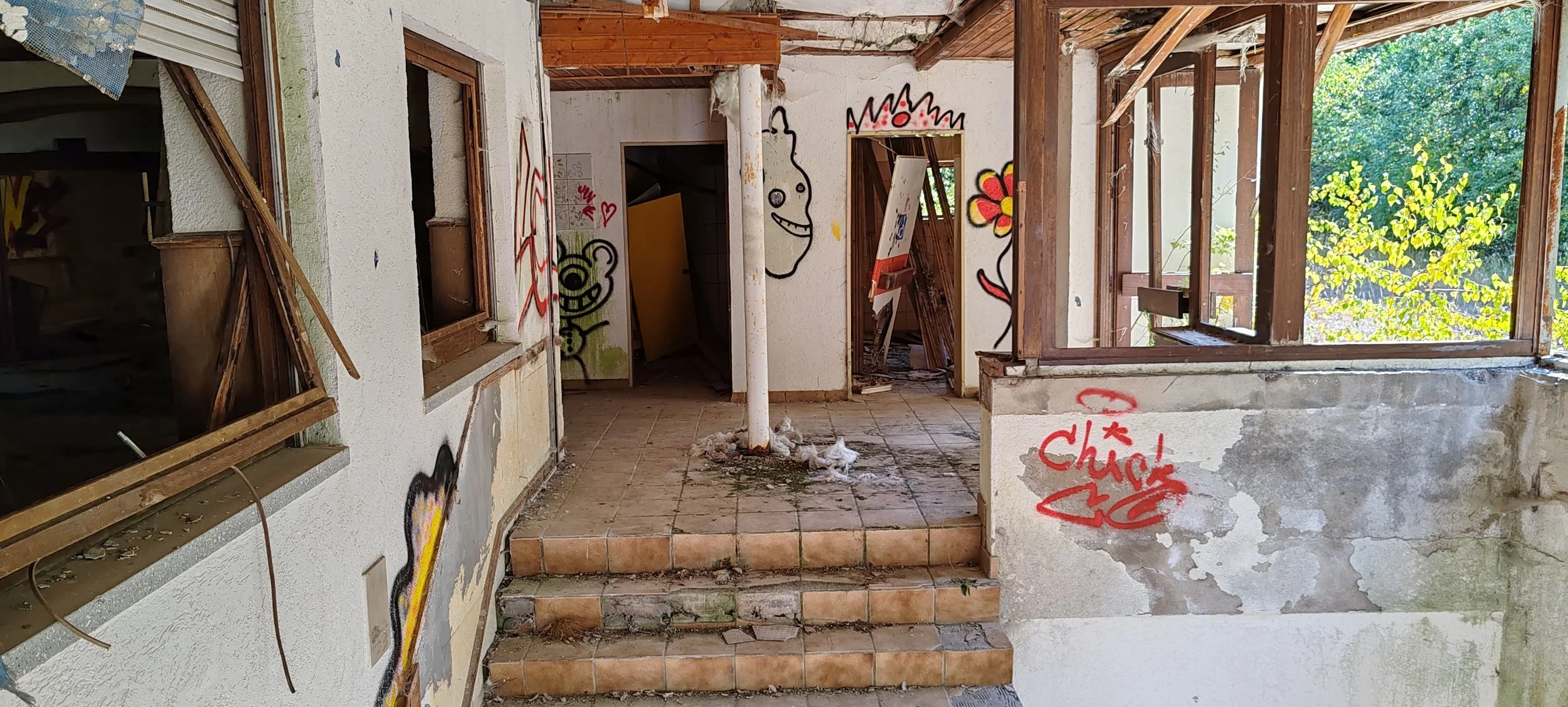 Verfallenes Haus mit Graffiti-Wandmalereien, offene Fenster, abgenutzte Treppen, und zerbrochenem Zustand, umgeben von grünen Bäumen.