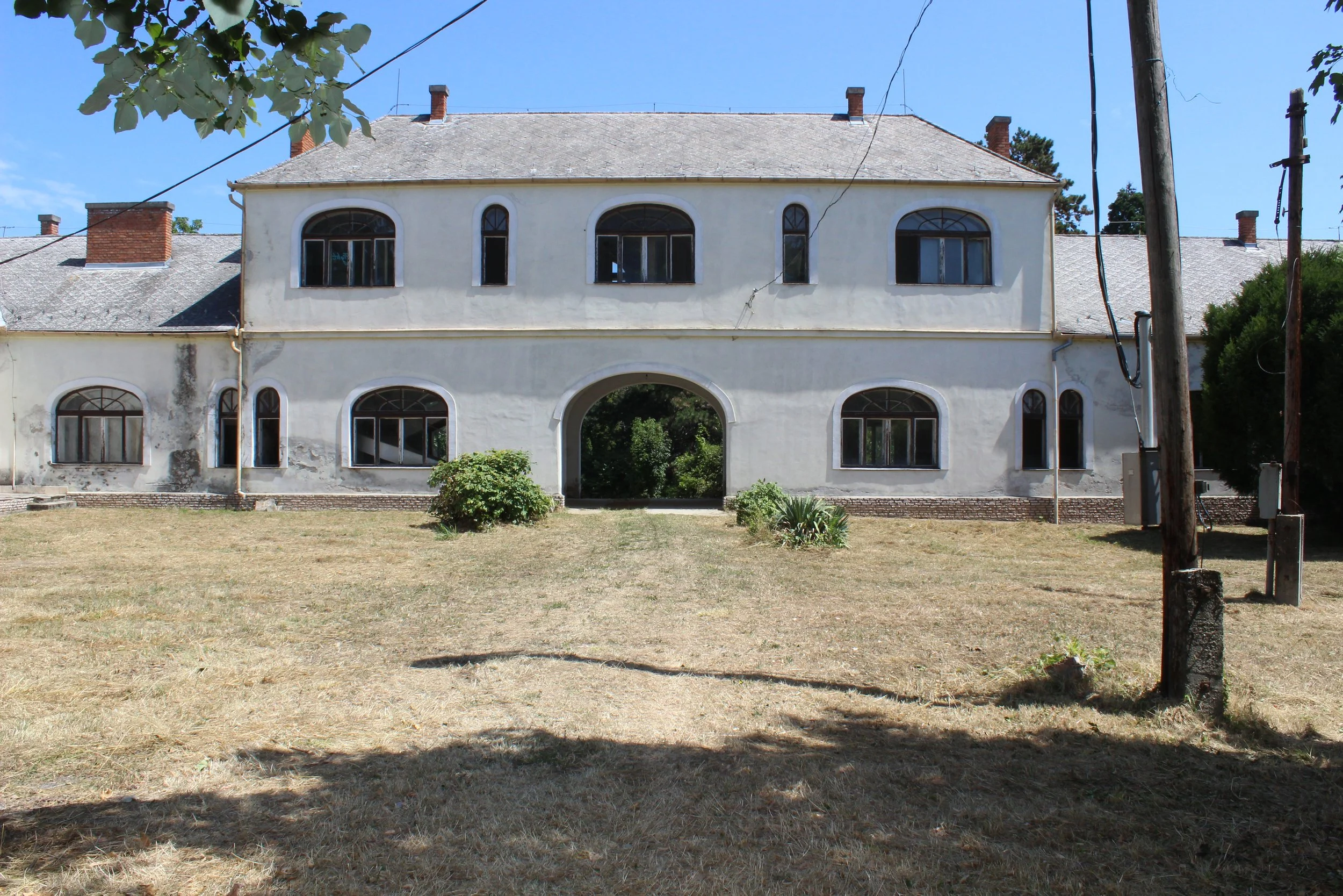 Ein altes, weißes, zweistöckiges Haus mit großen, arched Fenstern, einem großen Tor in der Mitte, umgeben von trockenem Gras und einigen Sträuchern, im Hintergrund ein blauer Himmel.