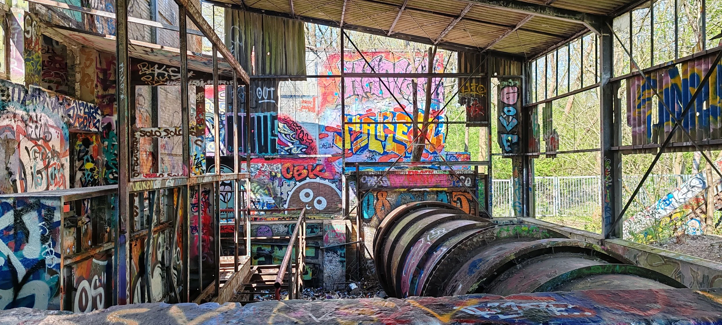 Verfallenes, mit Graffiti bedecktes Gebäude mit Metall- und Holzstrukturen im Inneren, umgeben von Bäumen und Felsen im Freien.