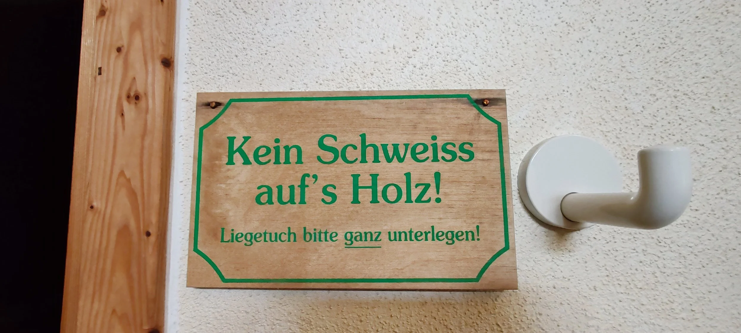 Holzschild mit grünem Rahmen und Text auf einer weißen Wand, der besagt: Kein Schweiss auf's Holz! Liegetuch bitte ganz unterlegen!