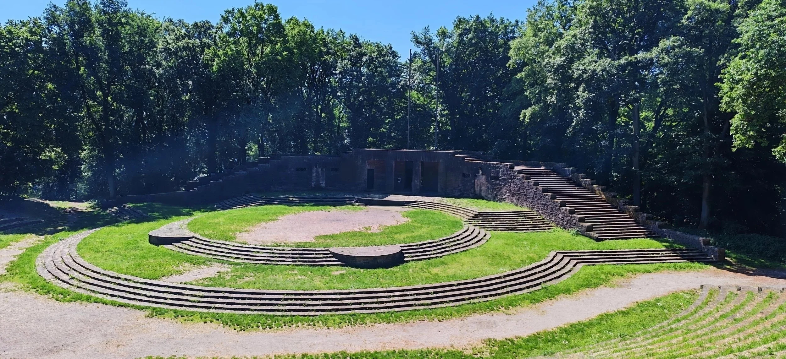 Ein antikes freiluft Theater mit halbkreisförmigen Steinstufen, umgeben von Bäumen und grünem Gras bei sonnigem Himmel.