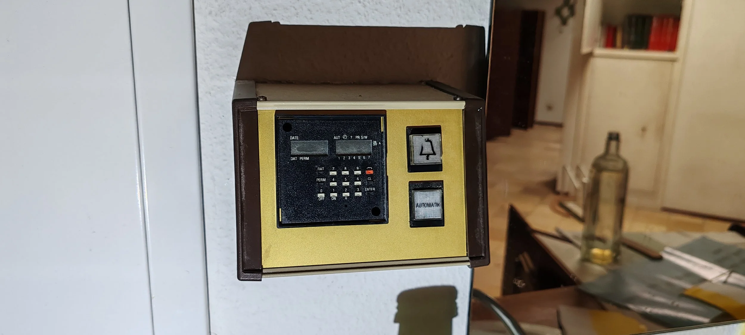 Alte elektronische Steuerungseinheit an einer Wand, mit schwarzem Bildschirm, Tasten und Schaltern, in einem Raum mit Büchern und Getränk im Hintergrund.