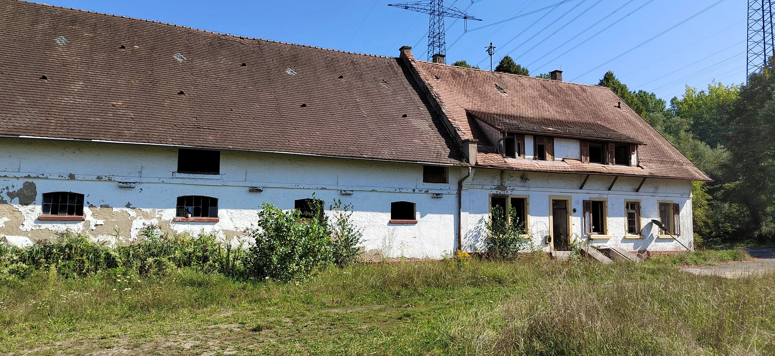 Ein altes, verlassenes Haus mit weißen Wänden, einigen Fenstern mit Holzläden und einem roten Ziegeldach, umgeben von Wildgras und Büschen, im Hintergrund einige Hochspannungsleitungen und eine bewaldete Hügelkette.