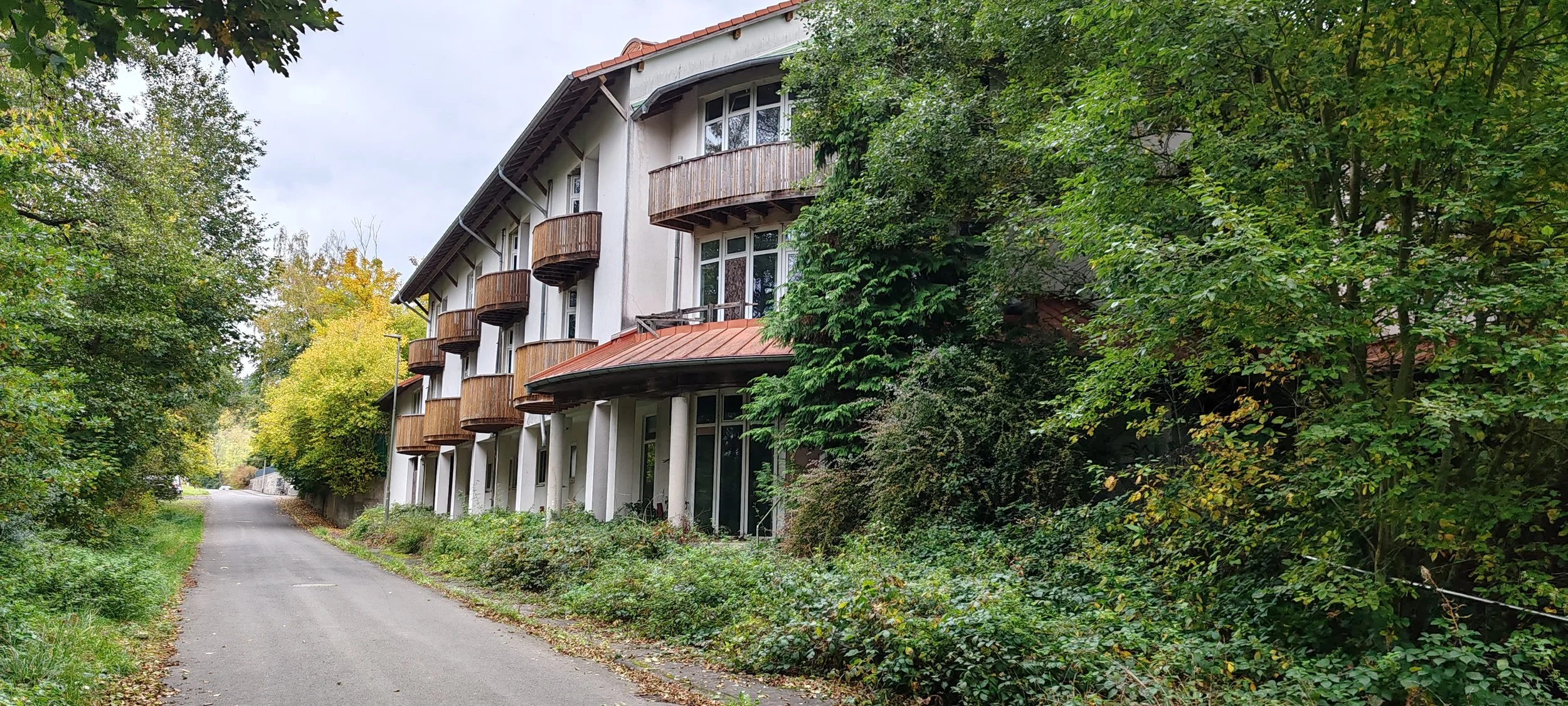 Mehrstöckiges Wohngebäude mit weißen Wänden, kleinen, runden Balkonen aus Holz, an einer geschwungenen Straße, umgeben von Bäumen und Grünflächen.