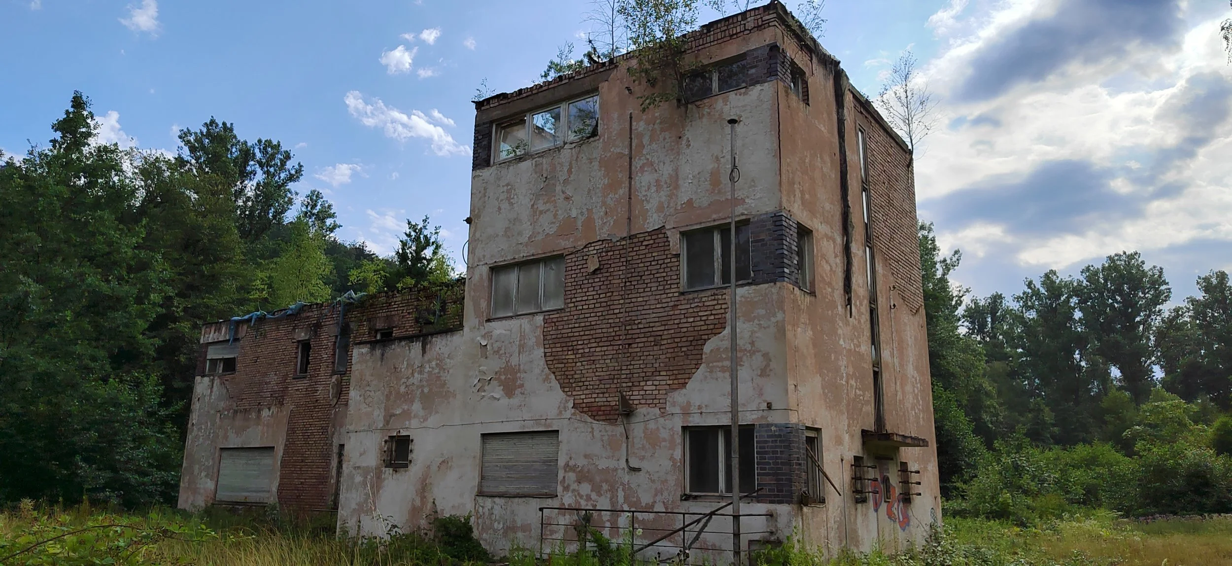 Verfallenes, mehrstöckiges Gebäude mit abblätternder Fassade und Graffiti an der Wand, umgeben von Grünflächen und Bäumen.