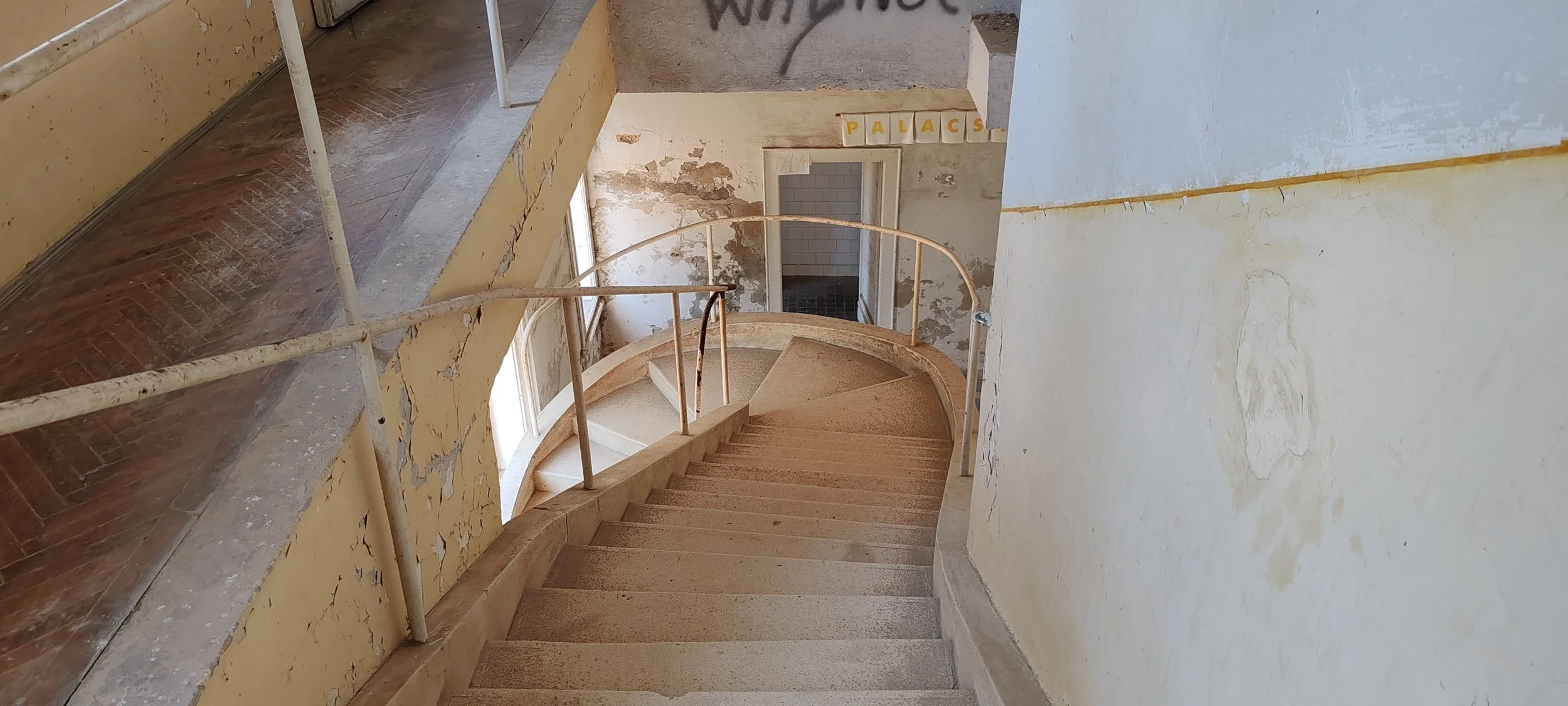 Ein veralteter, schmutziger, spiralförmiger Treppenaufgang in einem alten Gebäude mit beschädigten, streifigen Wänden, eines halb offenen Fensters und einem Raum mit Ziegelwänden im Hintergrund.