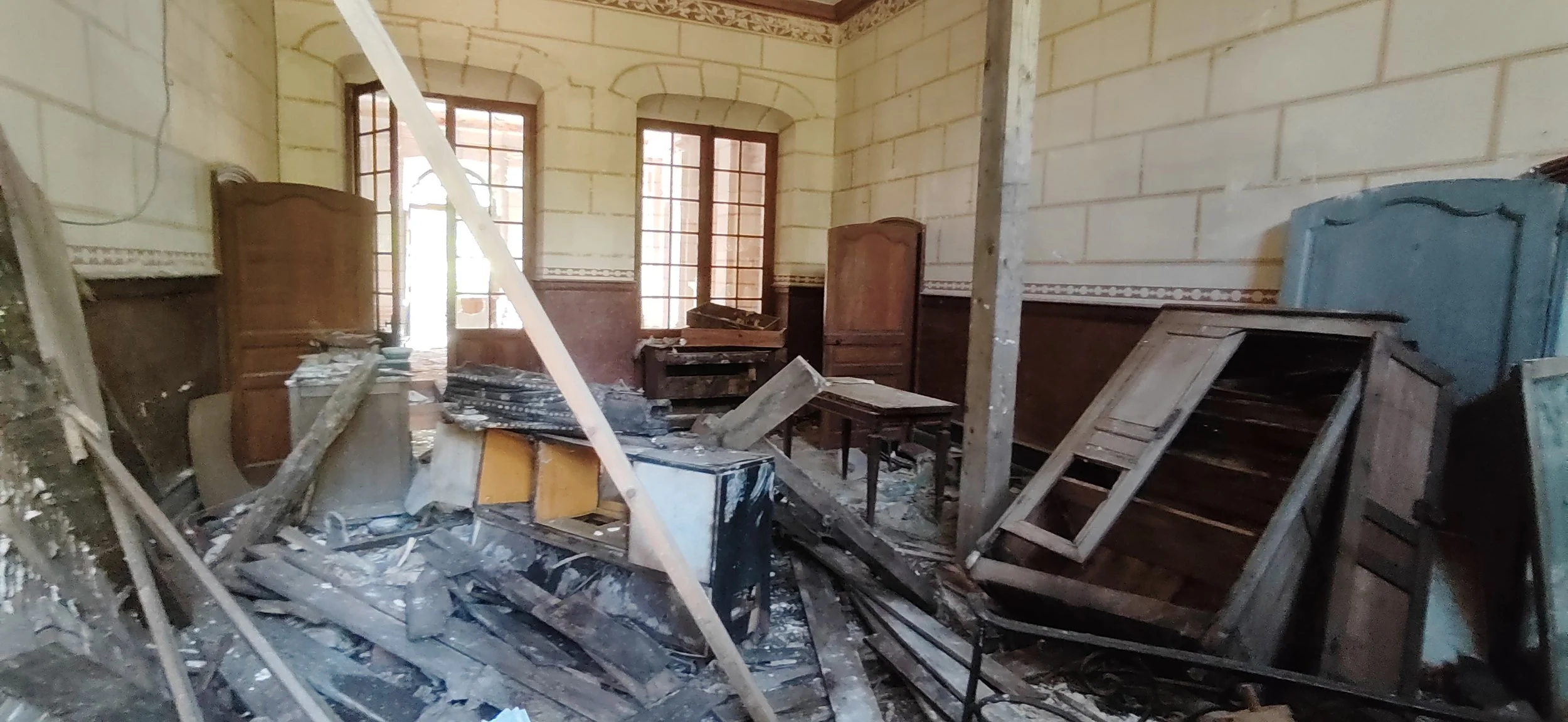 Ein Raum mit zerbrochenen Möbeln und zerklatschten Gegenständen, die Wände sind gelb mit braunen Ornamenten an der Decke, Fenster sind offen, es sieht aus wie ein Raum, der beschädigt wurde