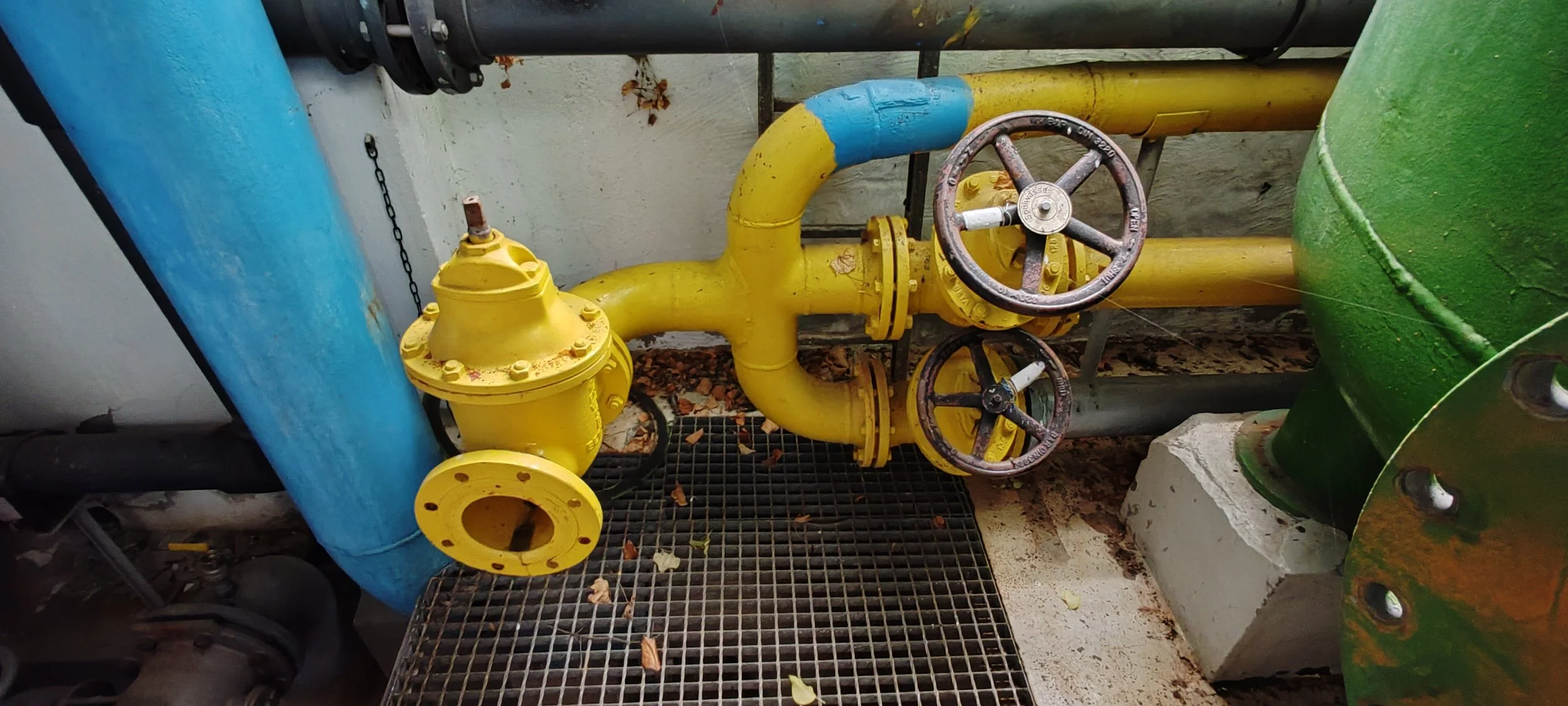Blick auf gelbe Wasserrohre mit Ventilen und zwei Griffen, verbunden mit einem blauen Rohr und einem grünen Behälter, in einer technischen oder industriellen Einrichtung.