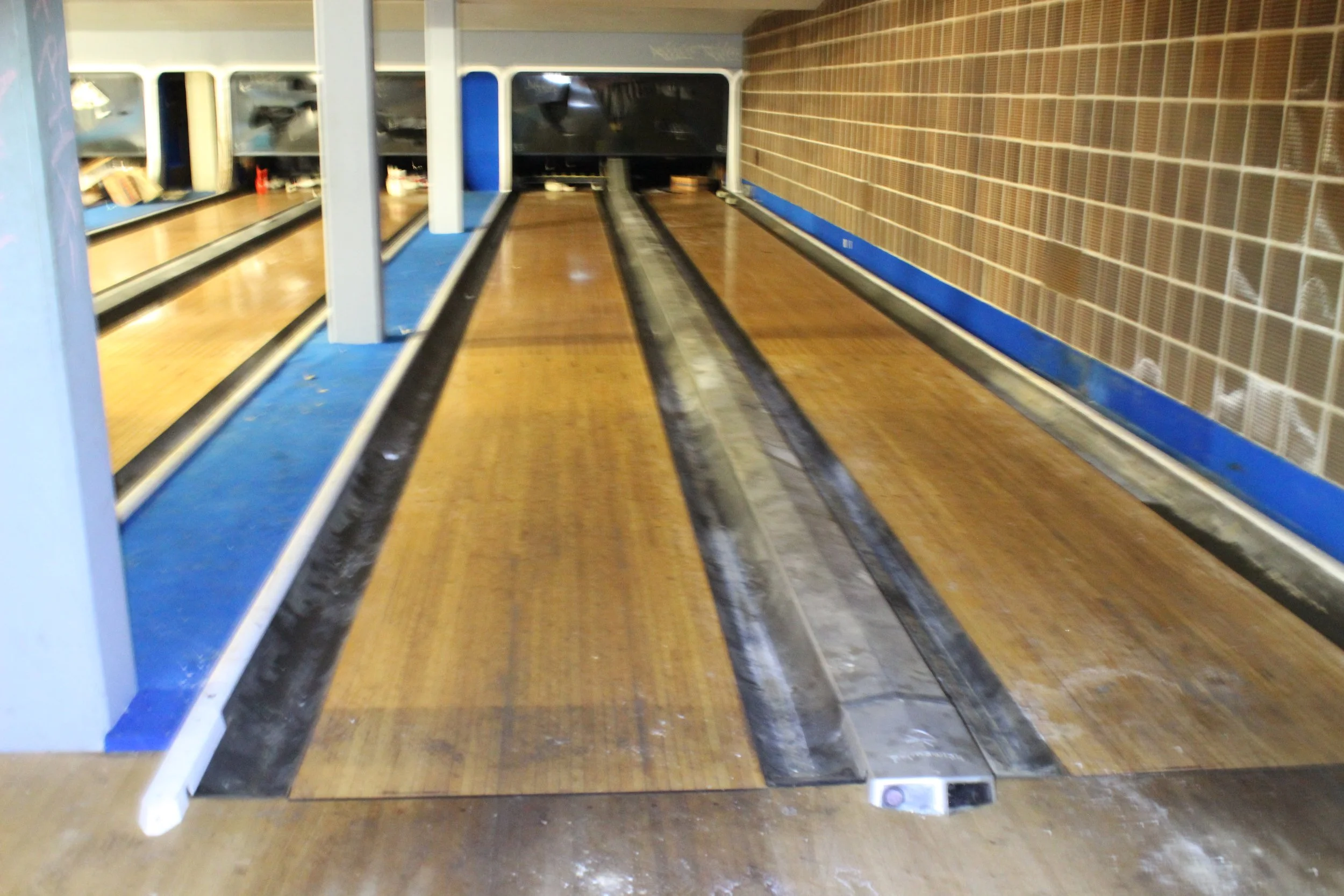 Leerer Bowlingbahnen mit Holzbelag und schwarzen Kanten, im Hintergrund die Bahnabschlusswand.