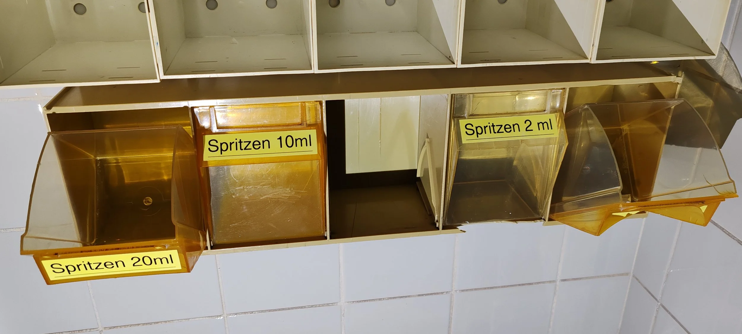 Aufbewahrungsboxen mit gelben Labels, die Spritzmengen von 10 ml, 2 ml und 20 ml anzeigen, auf einer Metallregal im Badezimmer.