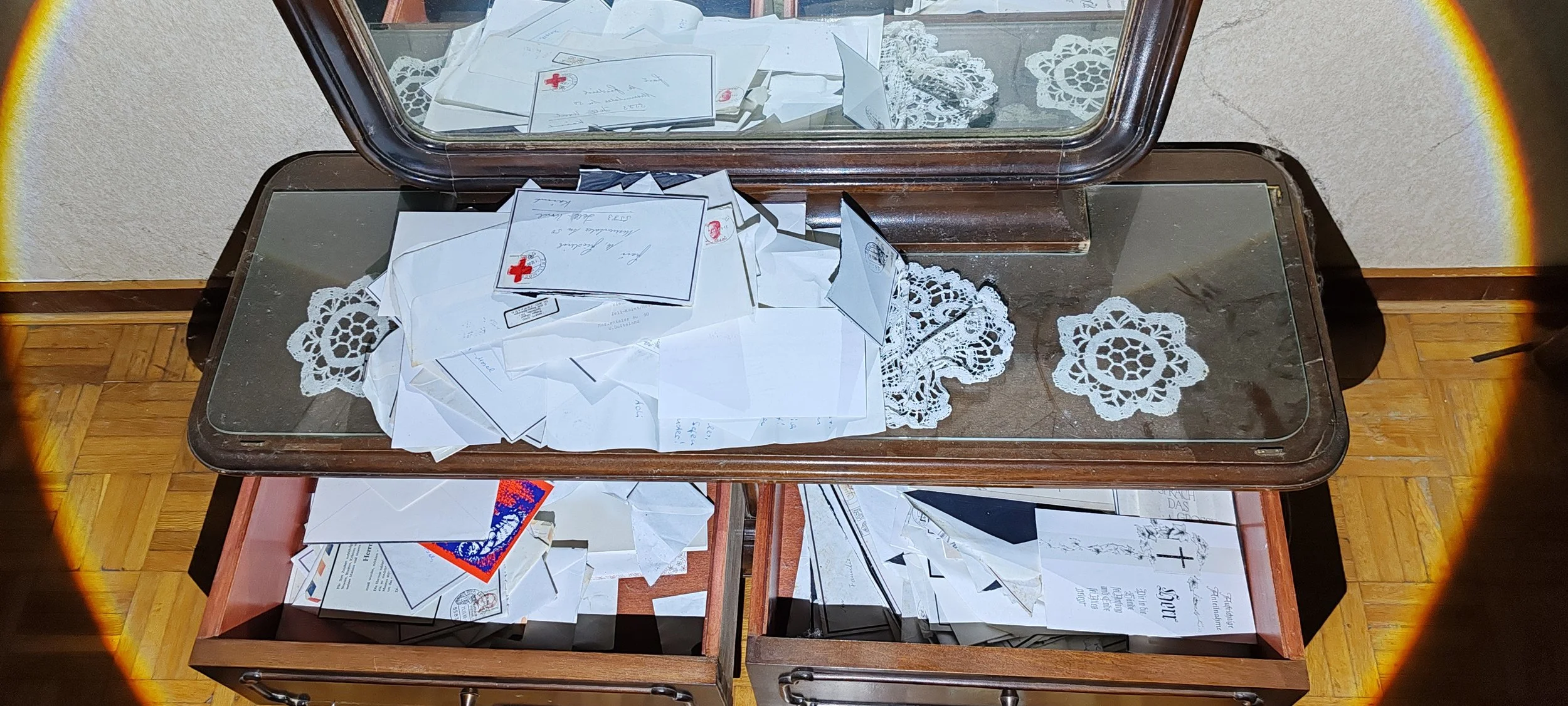 Ein Holztisch mit einer Glasscheibe darauf, die von einer Sockelglasplatte bedeckt ist und darauf liegen Briefe, Umschläge und Karten, darunter weiße doilies, in einem Raum mit Holzparkettboden.