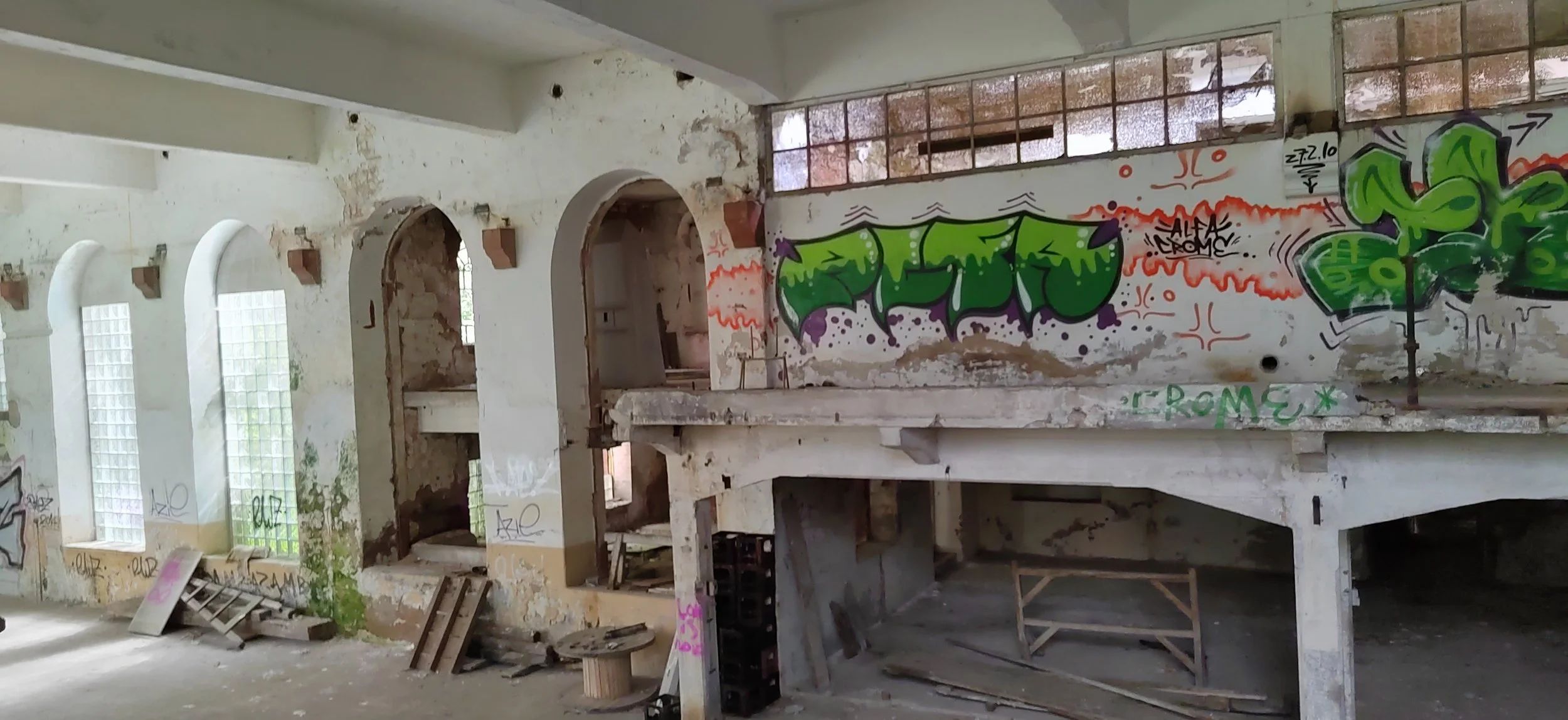 Verfallene, graffiti-bedeckte Innenraum eines Gebäudes mit hohen Fenstern, bröckelnden Wänden und Baustellenmaterialien auf dem Boden.