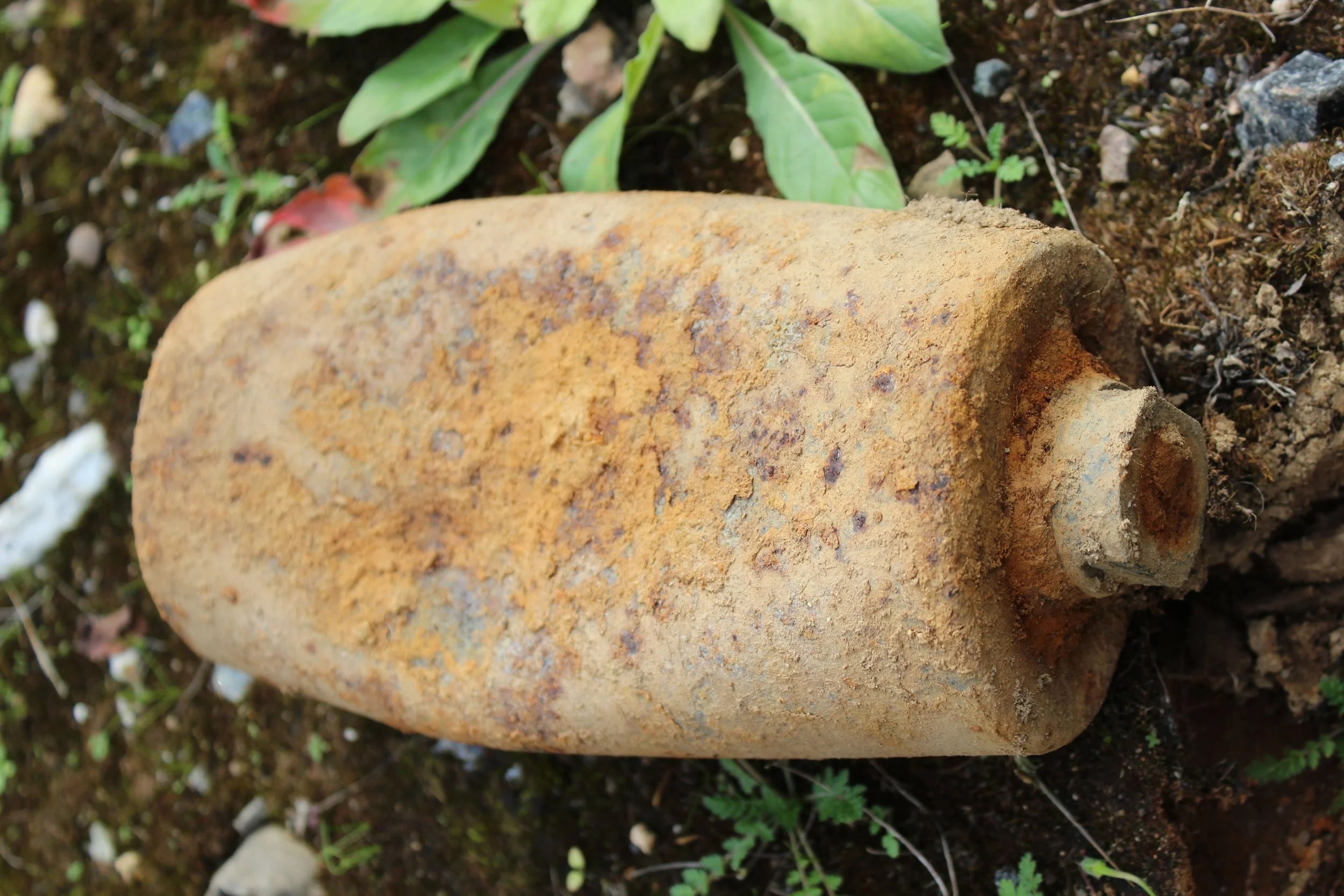 Ein verrosteter, rostfarbenen Munitionstyp im Boden liegend, umgeben von Erde, kleinen Steinen und Pflanzen.