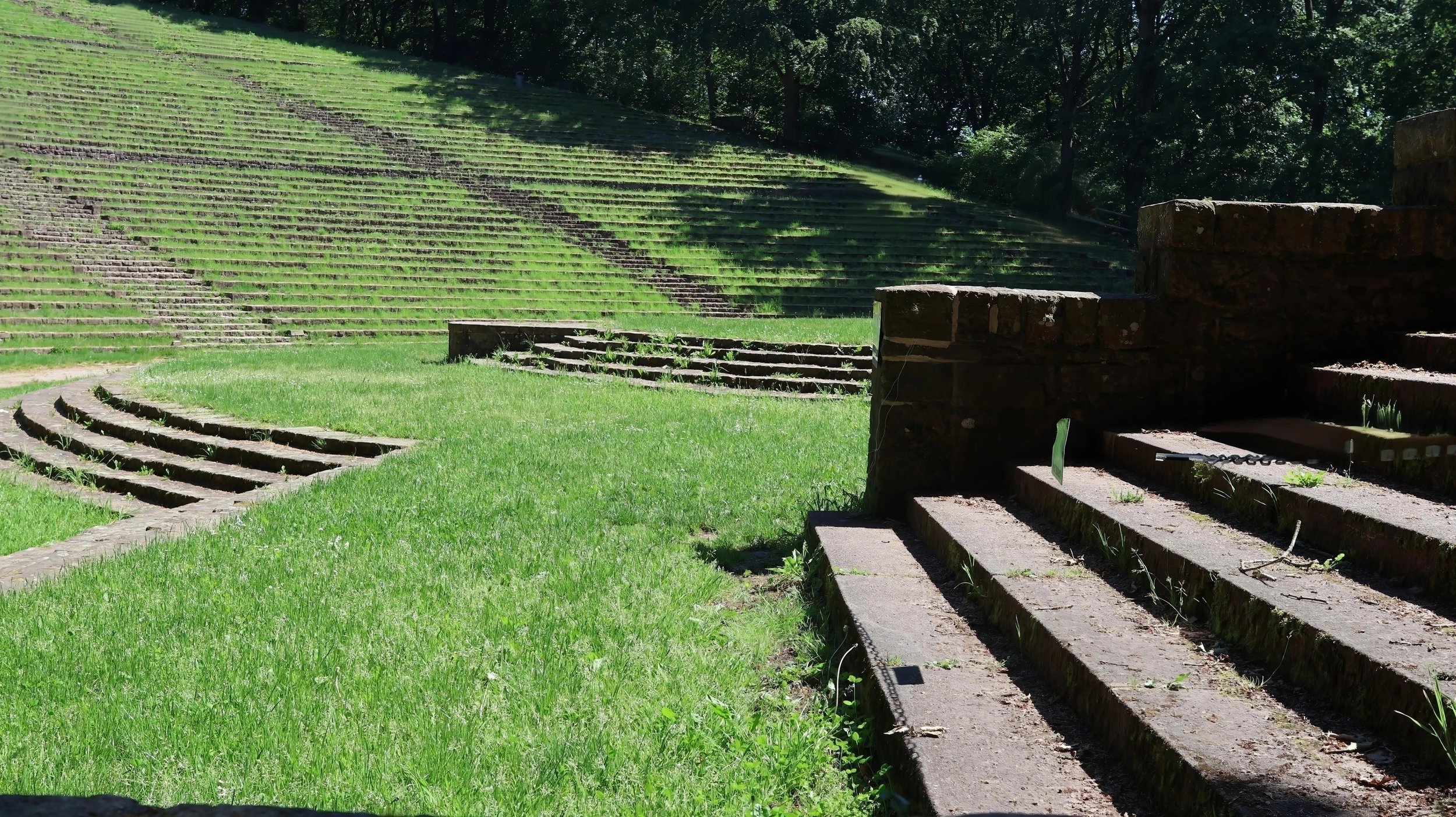 Altes Amphitheater aus Stein mit Stufen und Treppen, umgeben von grüner Wiese und Bäumen im Hintergrund.