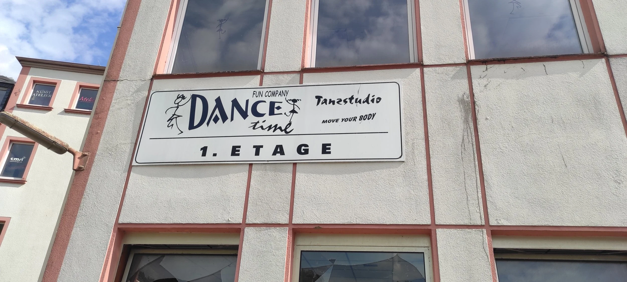 Schild an einem Gebäude mit Aufschrift 'Dance Time, Fun Company, Tanstudio, Move Your Body, 1. Etage' und Illustrationen von tanzenden Figuren.