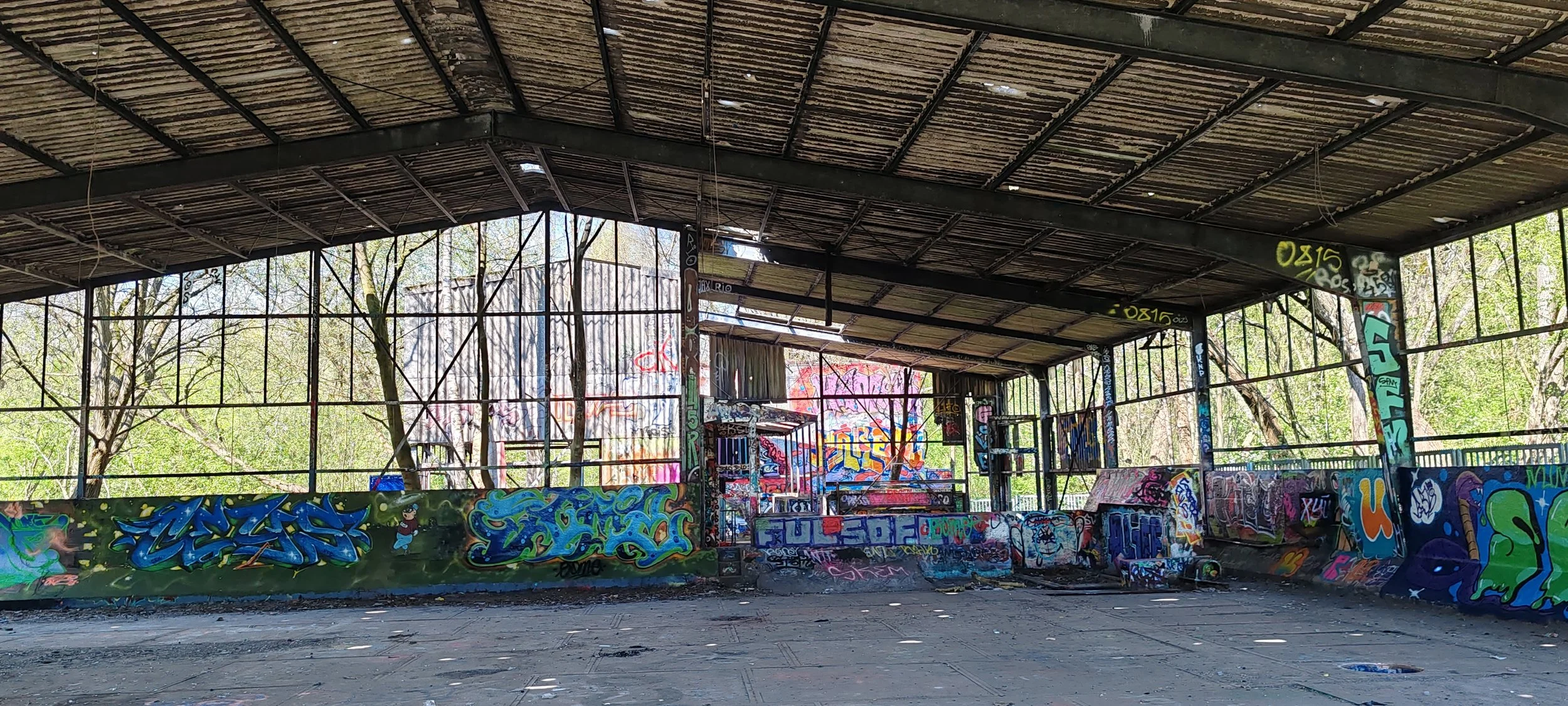 Verfallene, mit Graffiti bemalte Skatehalle im Freien, umgeben von Bäumen.