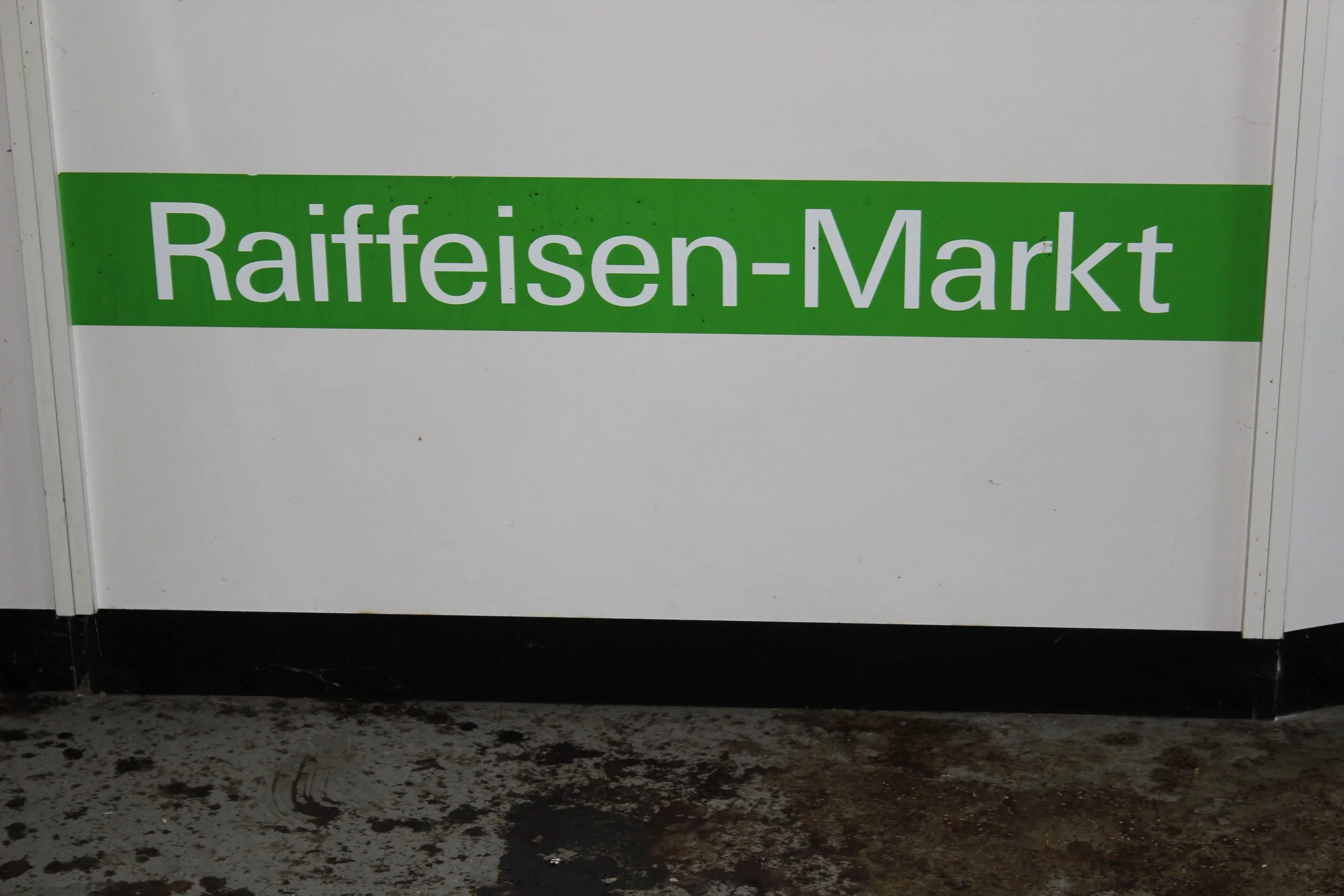 Grüner und weißer Schilder mit der Aufschrift 'Raiffeisen-Markt' an einer weißen Wand.