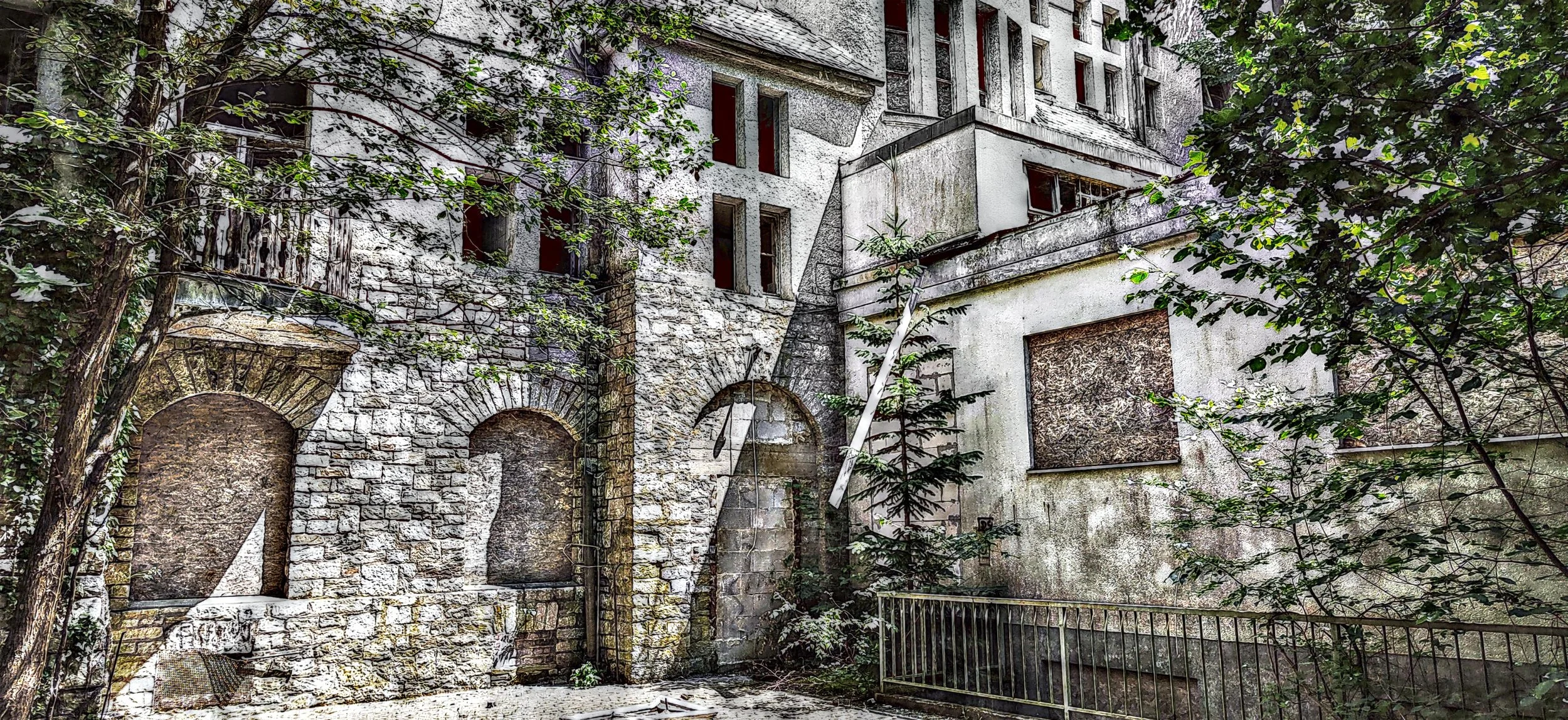 Verfallenes Gebäude mit zerbrochenen Fenstern und überwucherten Wänden, umgeben von Bäumen und Sträuchern.