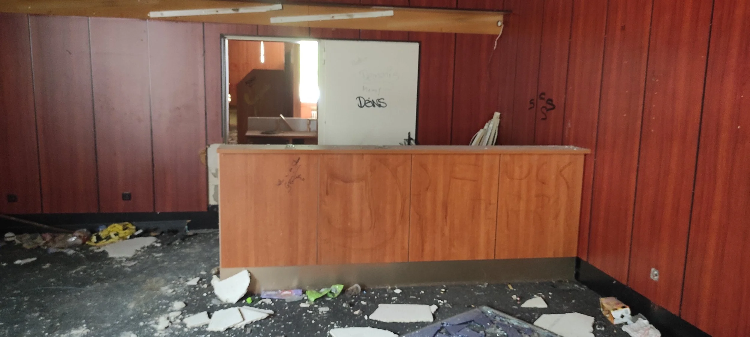 Innenaufnahme eines beschädigten Raumes mit einer Bar, Wänden in Holzoptik, verunreinigtem Boden, zerbrochenen Gegenständen und Graffiti an der Wand.