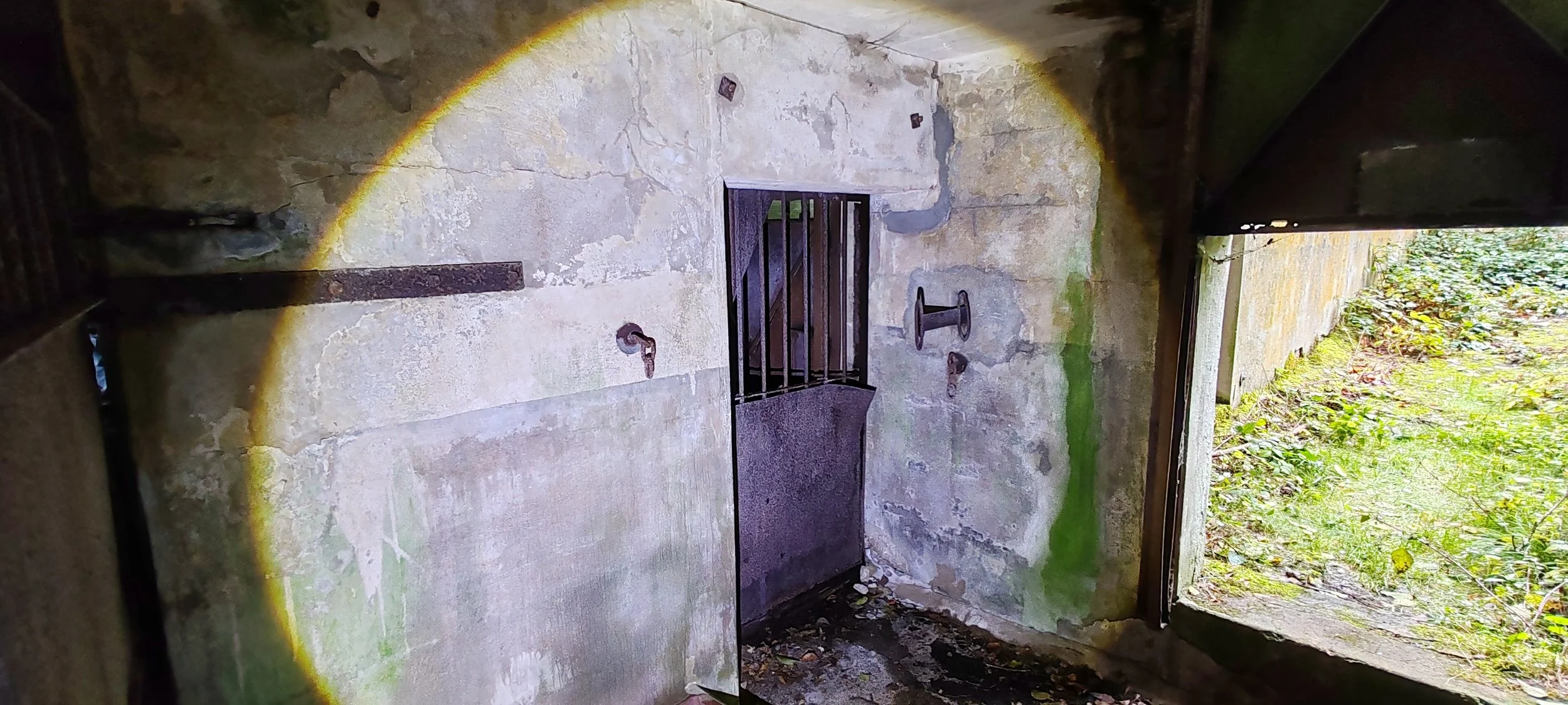 Ein altes, größtenteils verfallenes Gebäude mit einem offenen Fenster. Die Wände sind aus Stein und Beton, mit rostigen Metallarmaturen an der Wand. Im Außenbereich sieht man grünes Gras und Laub, das wahrscheinlich in einem Garten oder Wald liegt.