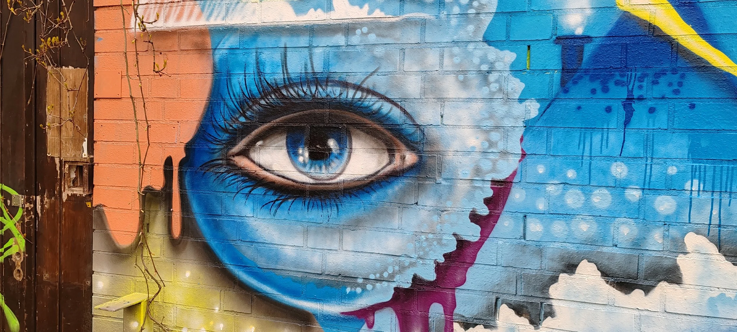 Graffiti-Malerei eines menschlichen Auges mit blauer Iris auf einer Mauer, umgeben von bunten Farbspritzern und Wolkenmotiven.