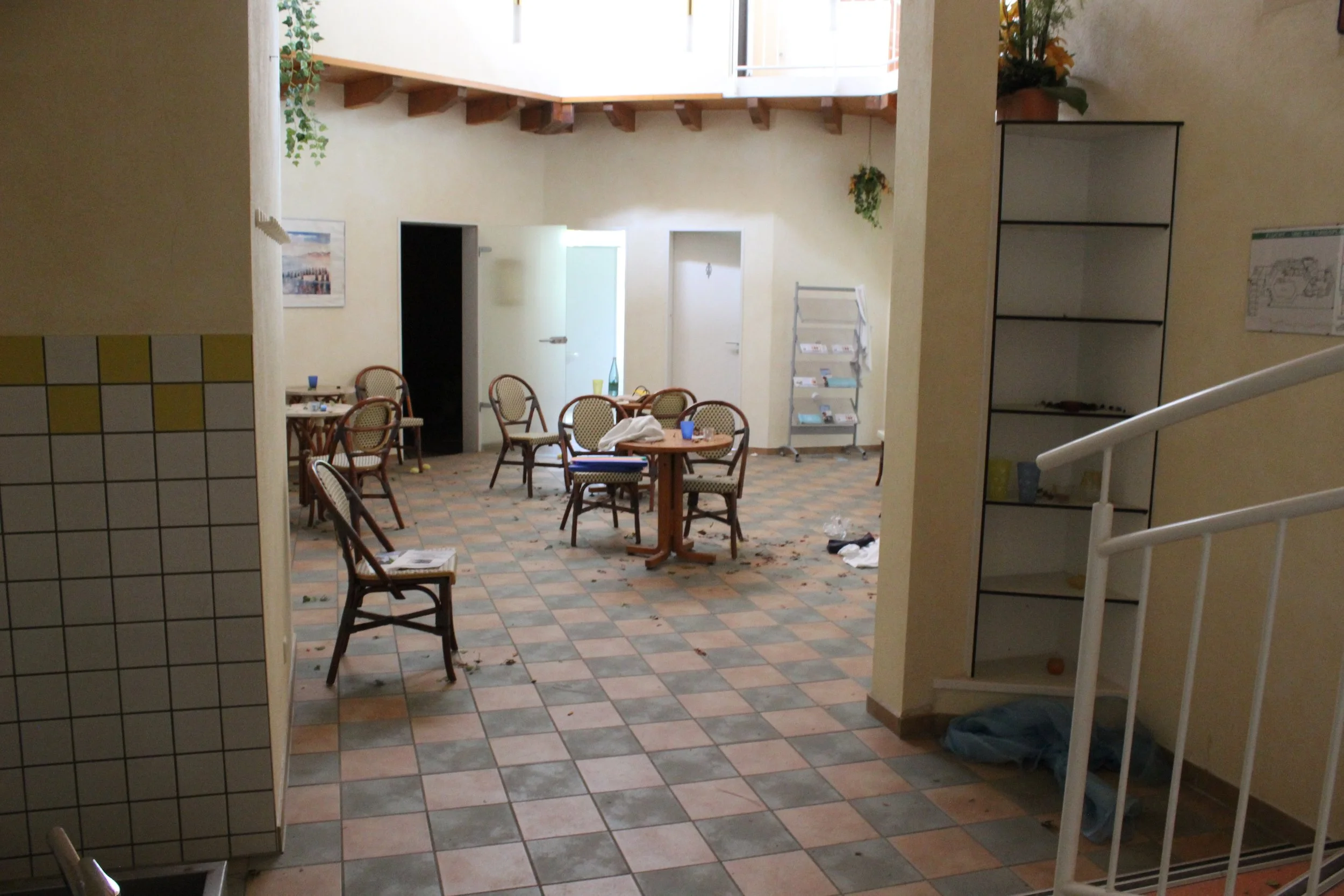 Unordentlicher Raum mit mehreren Stühlen und Tischen, einige mit Gegenständen wie Papieren, Flaschen und Tassen. Der Boden ist mit bunten Fliesen bedeckt, die Wände sind hellgelb. Es gibt eine offene Tür, eine Metallablage mit Zeitschriften und eine 