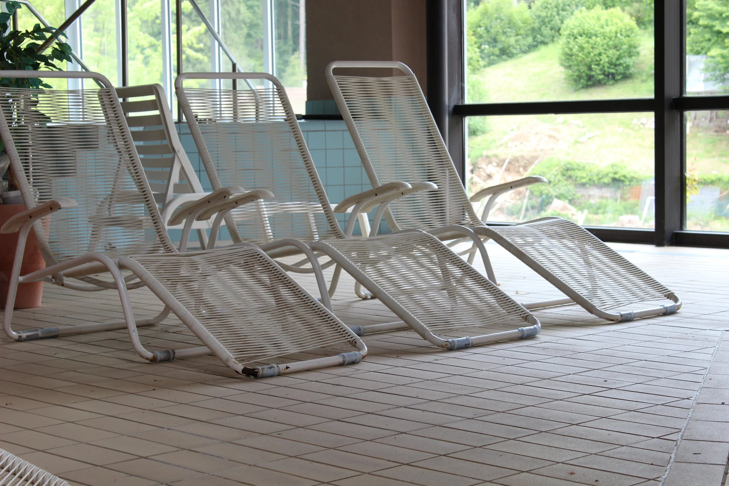 Leere Liegestühle in einem Spa-Booloop-Bereich mit großen Fenstern, durch die grüner Garten zu sehen ist.