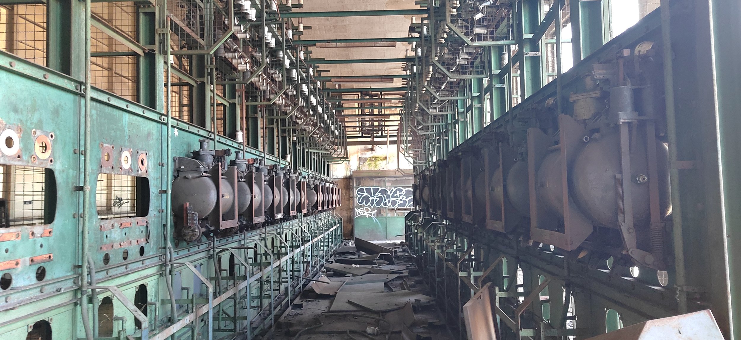 Verlassene Industrieanlage mit Metallstrukturen und Maschinen, graffiti an der Wand im Hintergrund, auf dem Boden liegen Trümmer und Gerümpel.