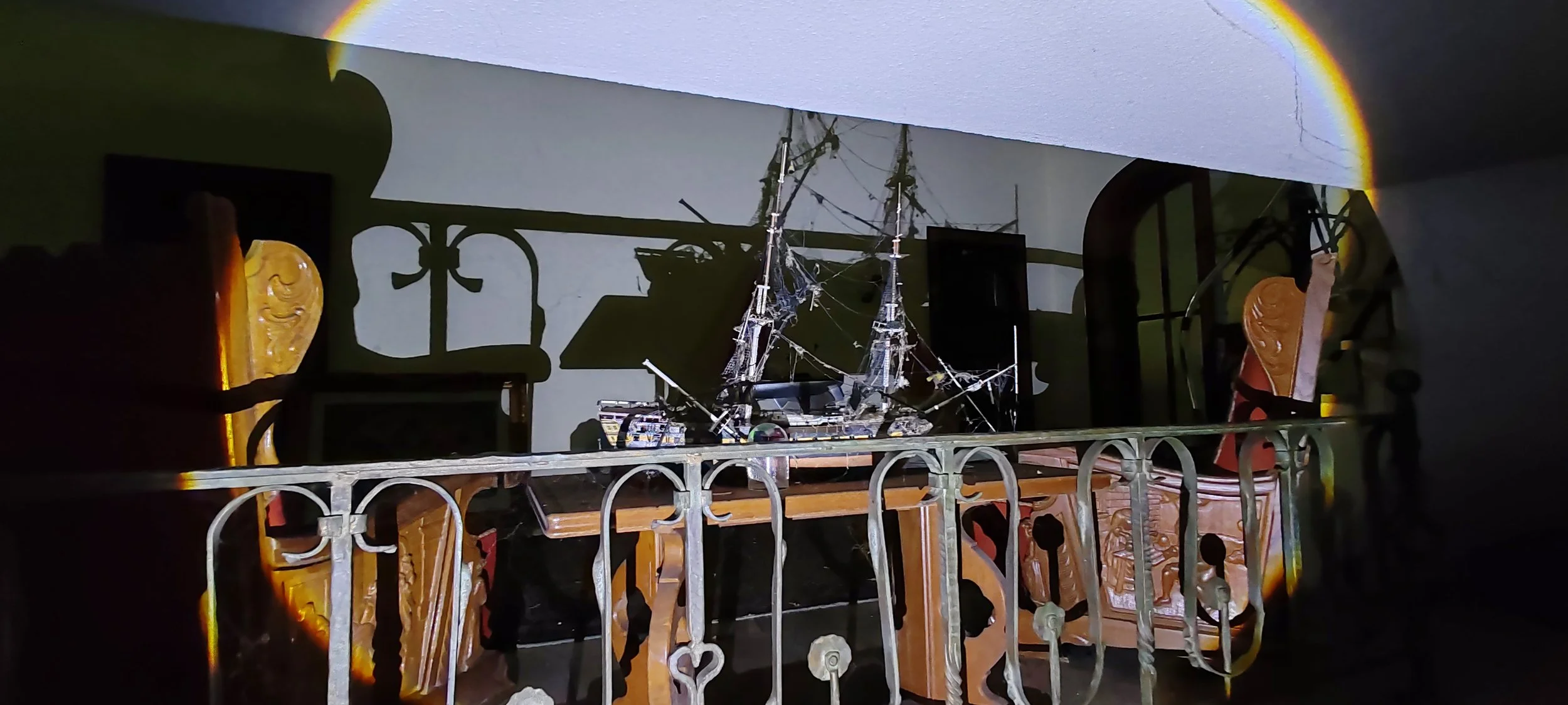 Ein detailliertes Modell eines Segelschiffes auf einem Tisch, umgeben von alten Holzstühlen, mit einer Lichtprojektion, die einen Regenbogen auf die Wand wirft.