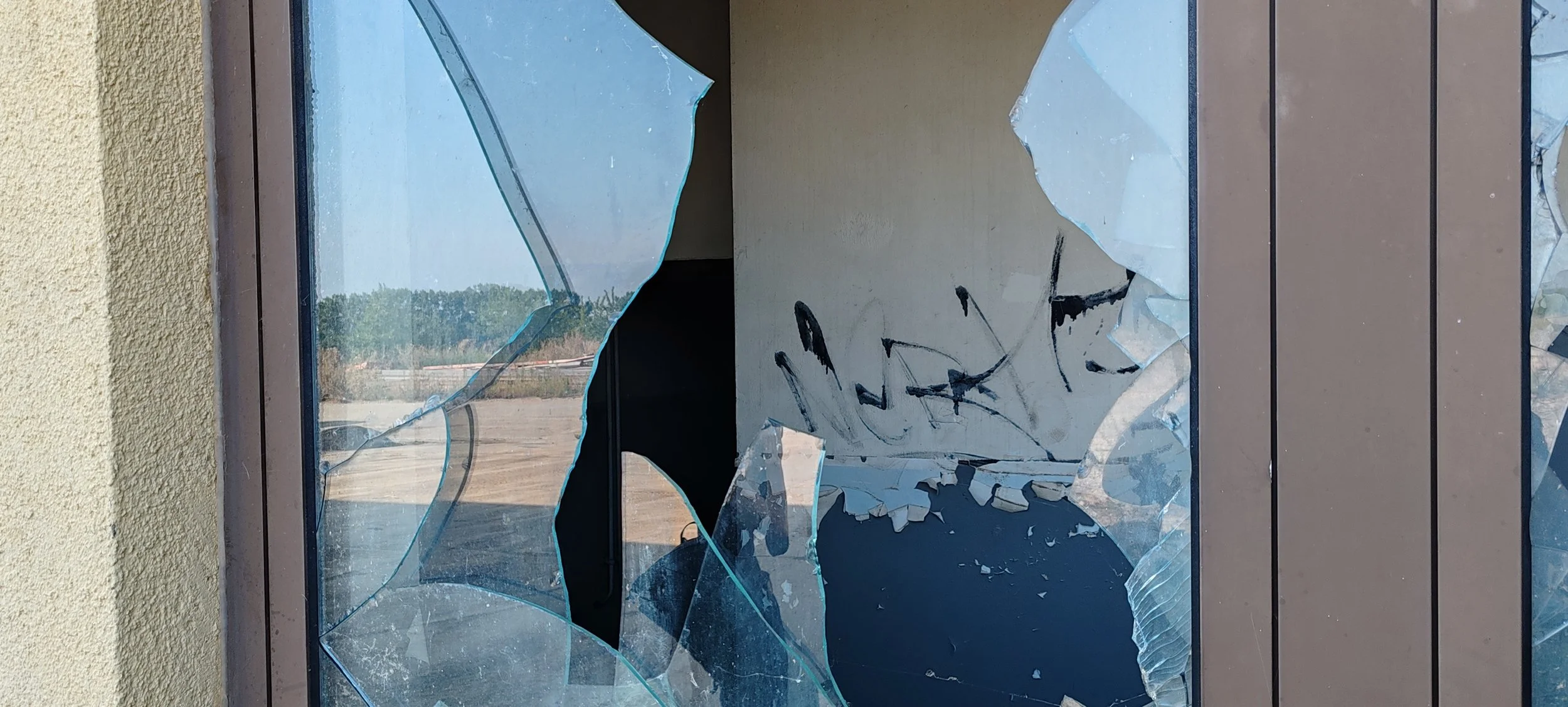 Ein zerbrochenes Fensterscheibe mit mehreren Rissen und Scherben. Im Hintergrund sind Bäume und ein offener Himmel sichtbar.