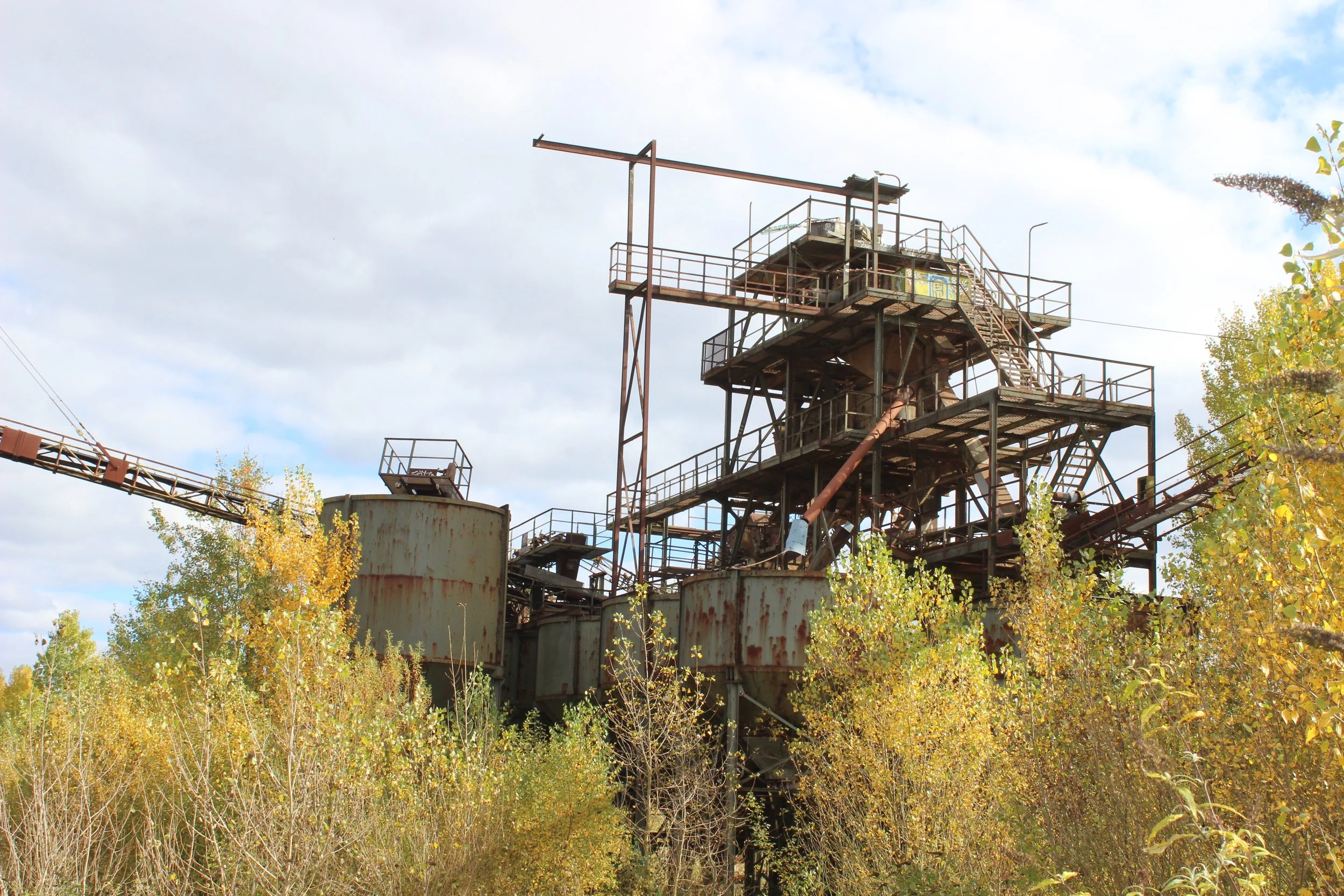 Verrostete, alte Industrieanlage im Freien umgeben von Büschen und Bäumen mit gelbgrünen Blättern, Himmel mit Wolken im Hintergrund.