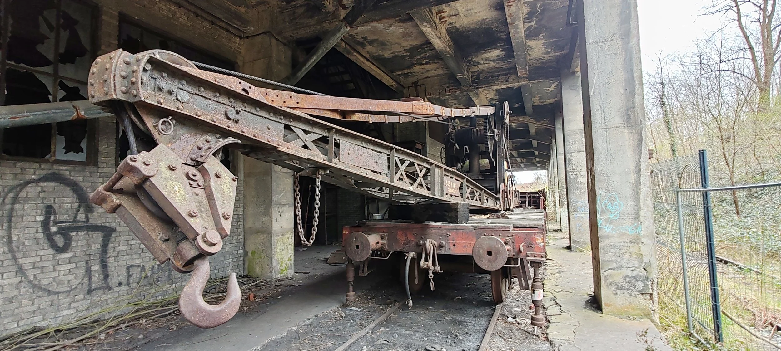 Ein alter, rostiger Kran auf Gleisen vor einer verfallenen, mit Graffiti besprühten Backsteinmauer und einer Brücke. Im Hintergrund sind Bäume zu sehen.