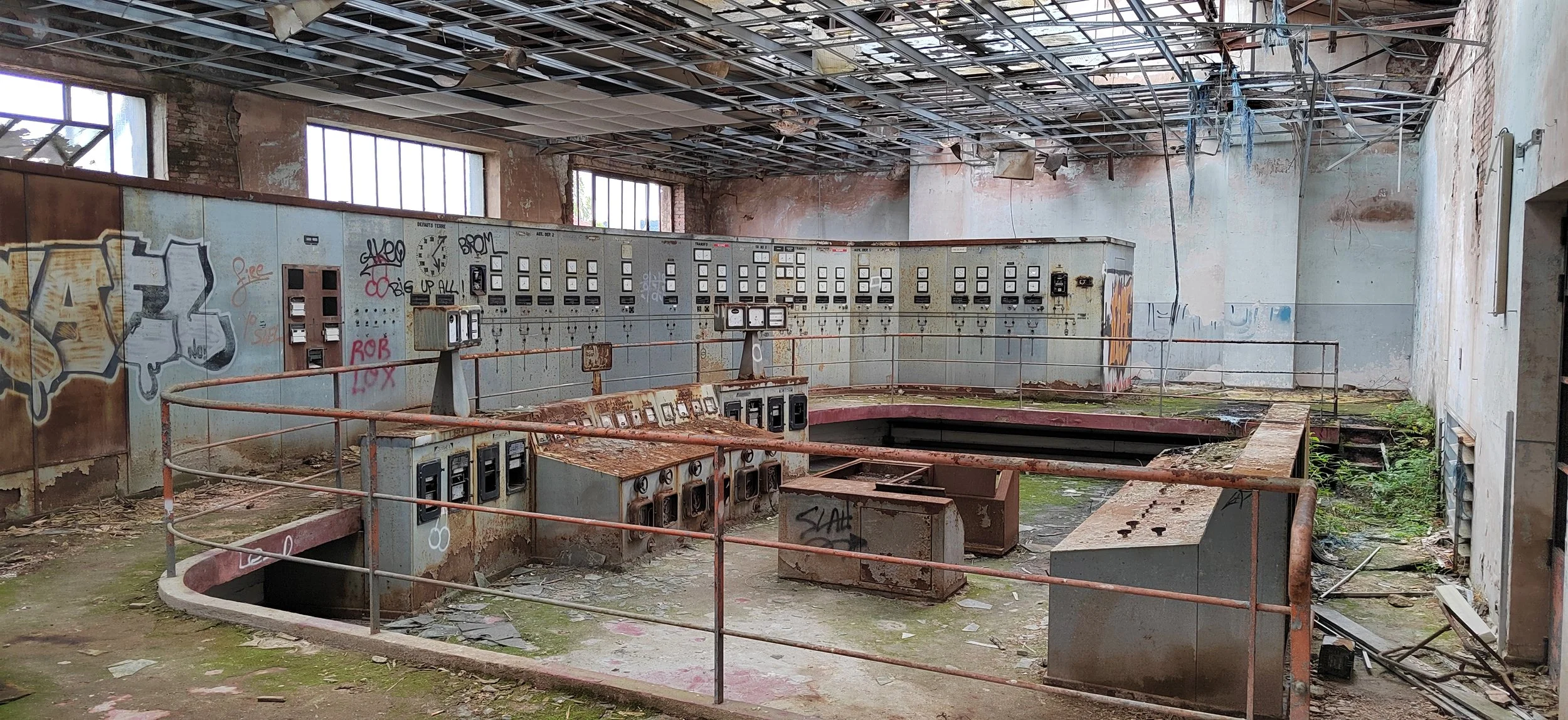 Verlassene, verrostete Kontrollzentrale in einem Raum mit zerbrochenen Fenstern, Graffiti und kaputtem Boden.