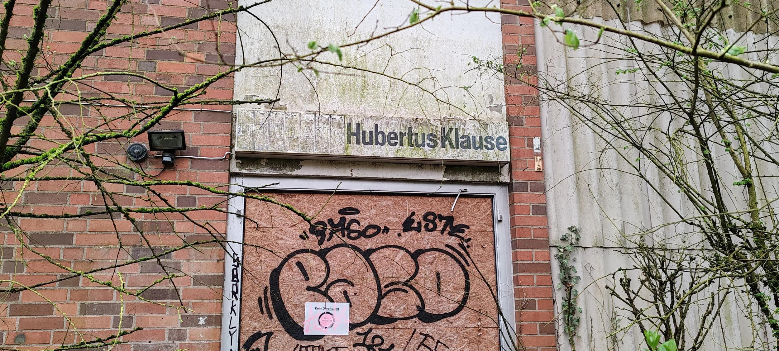 Verfallene Tür an einem ummauerten Gebäude mit Graffiti, umgeben von dünnen, blattlosen Ästen.